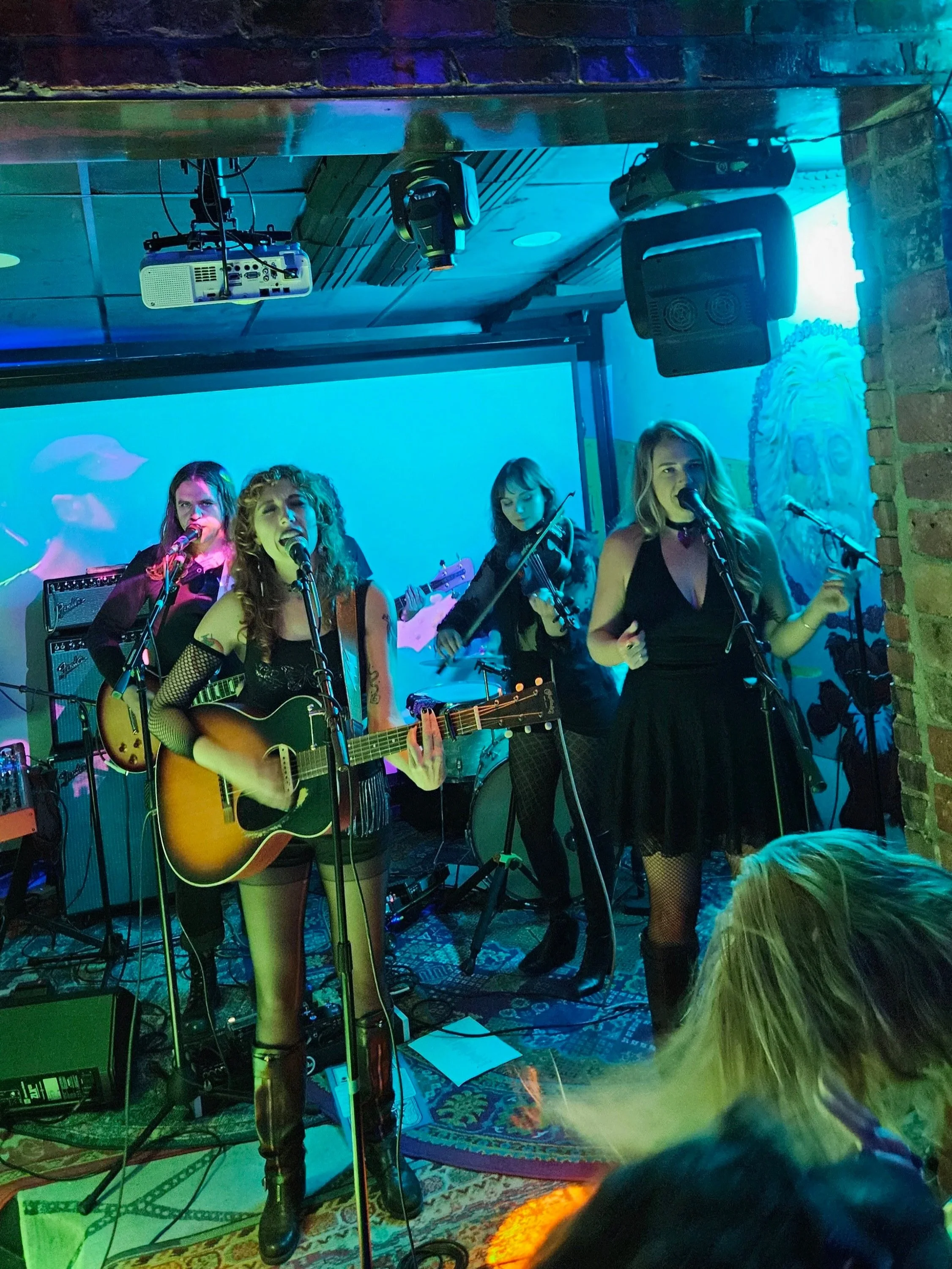 A band of five female musicians performing on stage in a dimly-lit venue, with one playing acoustic guitar and singing, and others playing drums, violin, and singing into microphones.