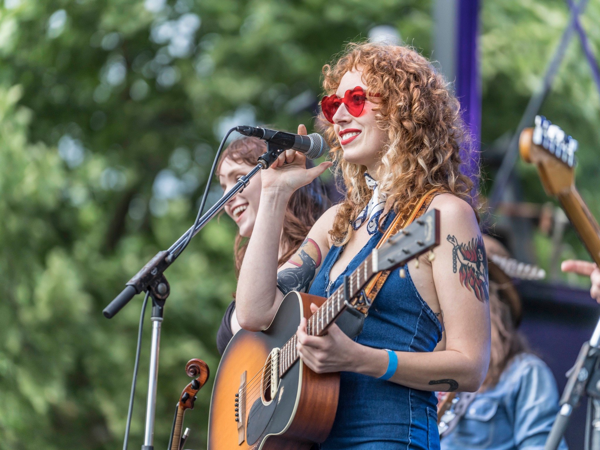 A woman with curly hair wearing red heart-shaped sunglasses plays an acoustic guitar and sings into a microphone during an outdoor performance, with another woman smiling in the background.