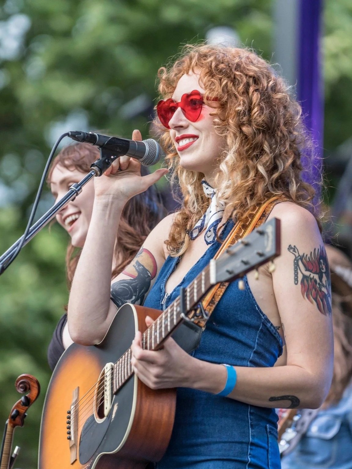A woman with curly red hair and sunglasses singing into a microphone while holding an acoustic guitar. She is wearing a denim sleeveless top, has tattoos on her arms, and is performing outdoors with a blurred green background.