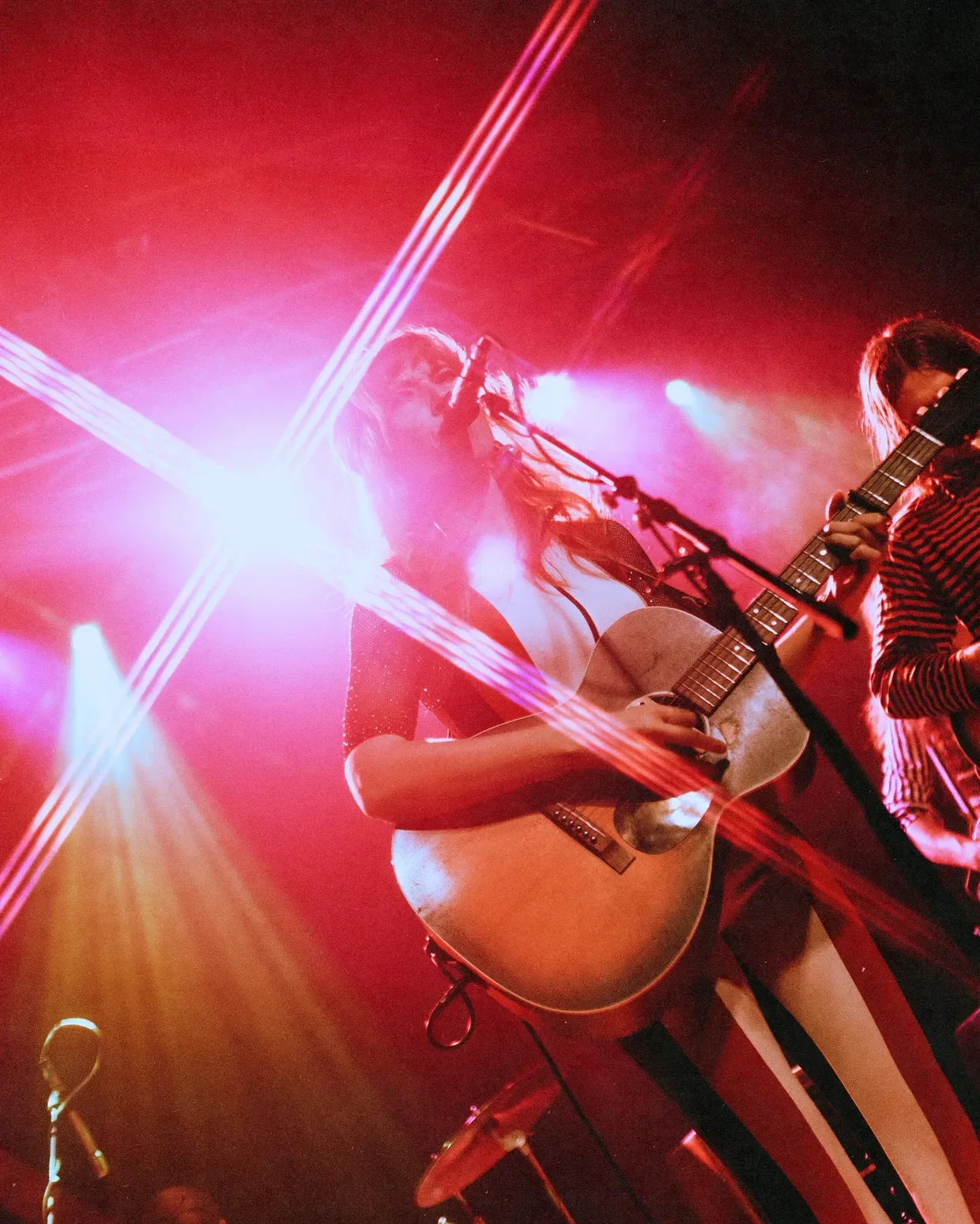 A female musician playing an acoustic guitar on stage with pink and purple lighting and laser beams.
