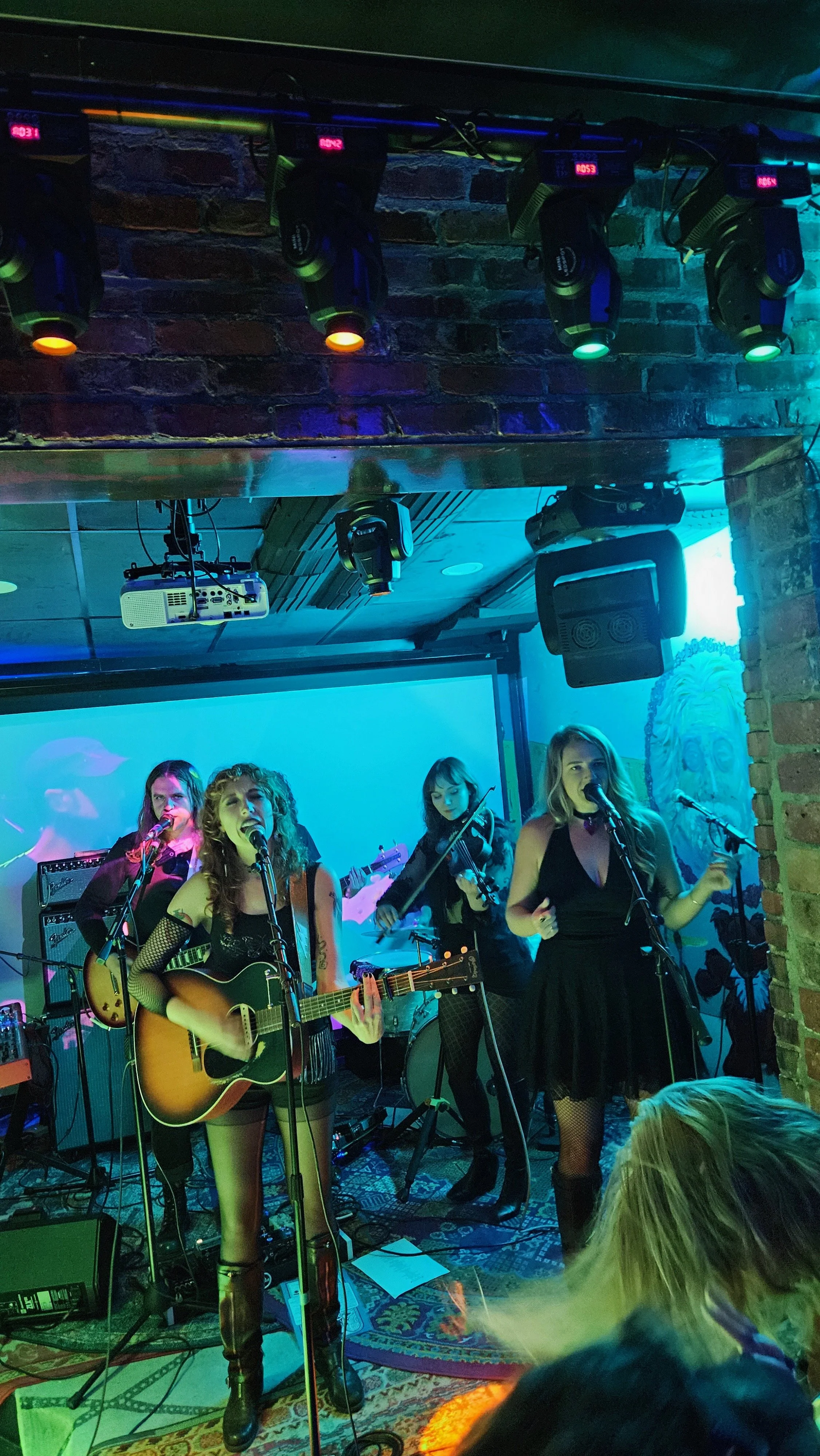 A live band performance in a dimly lit venue with brick walls, featuring four female musicians playing guitars and singing, with a large screen in the background and stage lighting overhead.