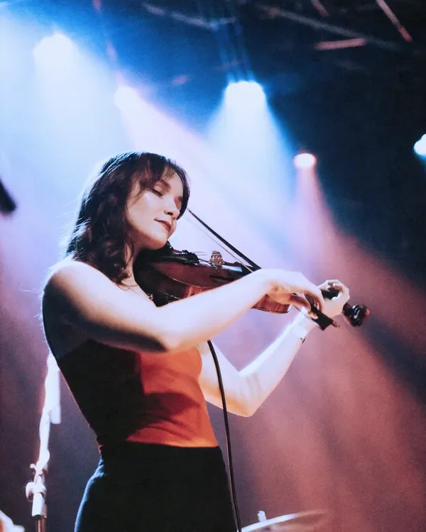 A woman playing the violin on stage under blue and pink concert lighting.