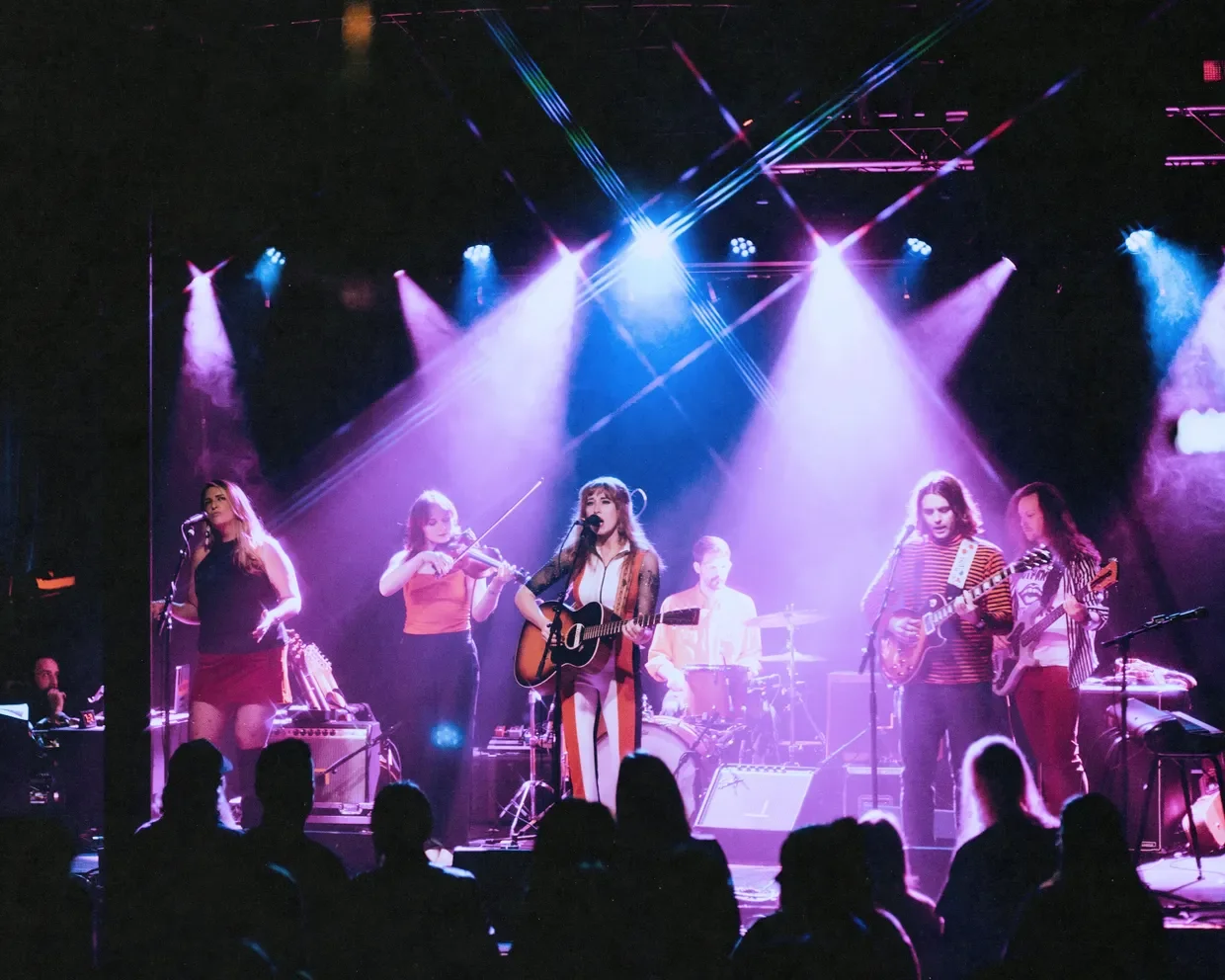 A musical band performing on a stage under colorful lights, with seven musicians including a singer playing guitar, violin, drums, and other instruments, and an audience watching.