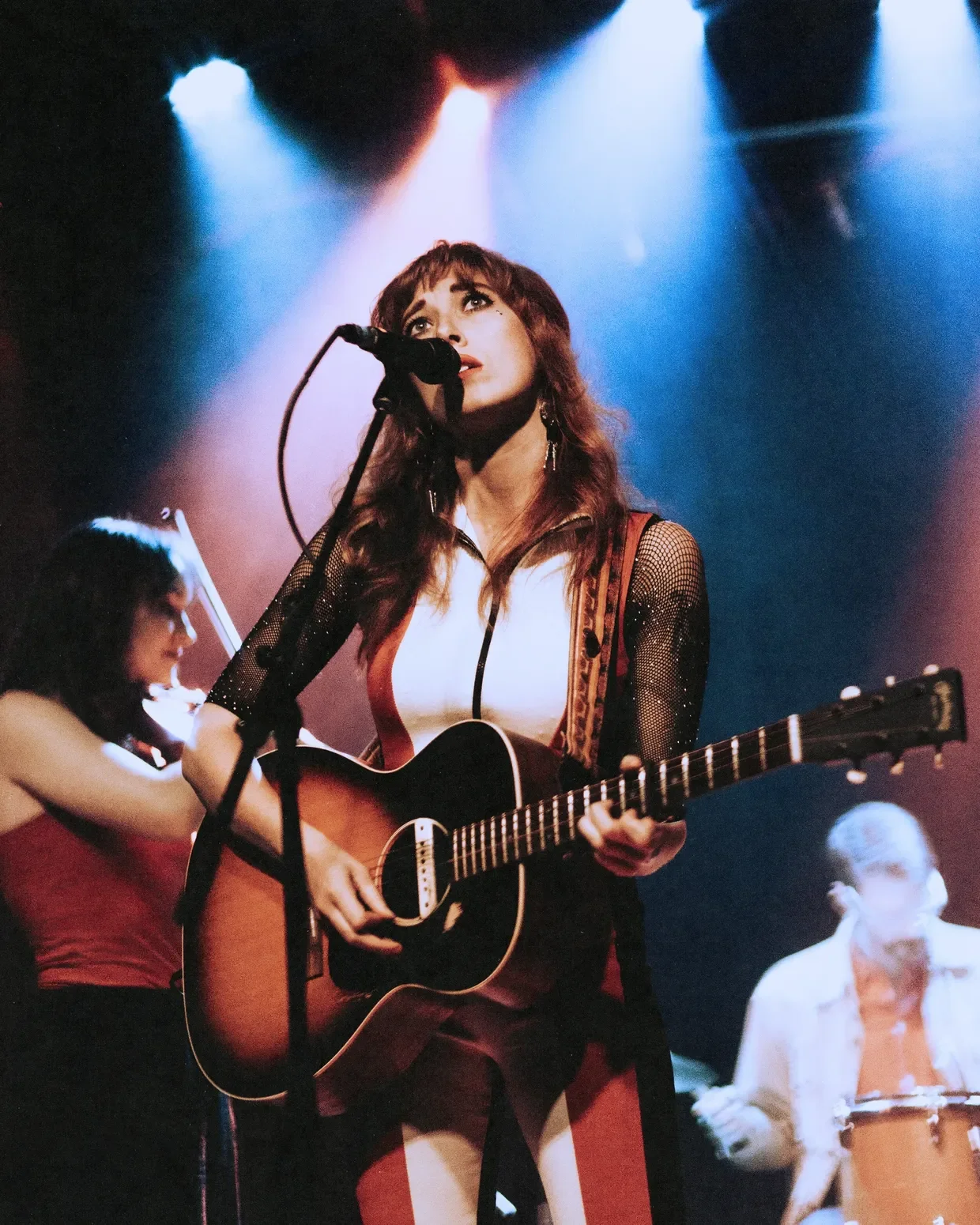 A female musician with long brown hair sings into a microphone while playing an acoustic guitar on stage, with colorful stage lights shining above her.