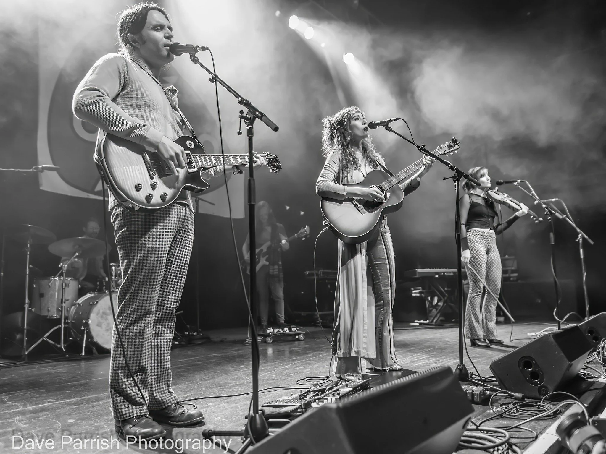 Black and white photo of three women performing on stage with guitars, singing into microphones, with a drummer in the background, stage lights and smoke effects.