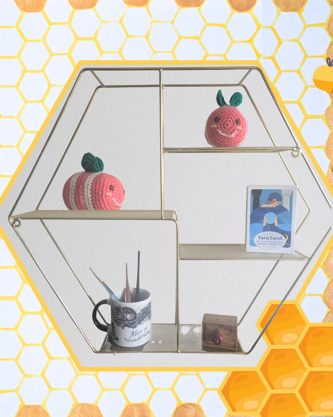 On a honeycomb shelf there is a pink and white amigurumi bee with green wings. There is a Yarn Tarot deck, measuring tape box, and Alice in Wonderland mug holding a paintbrush and crochet hooks.