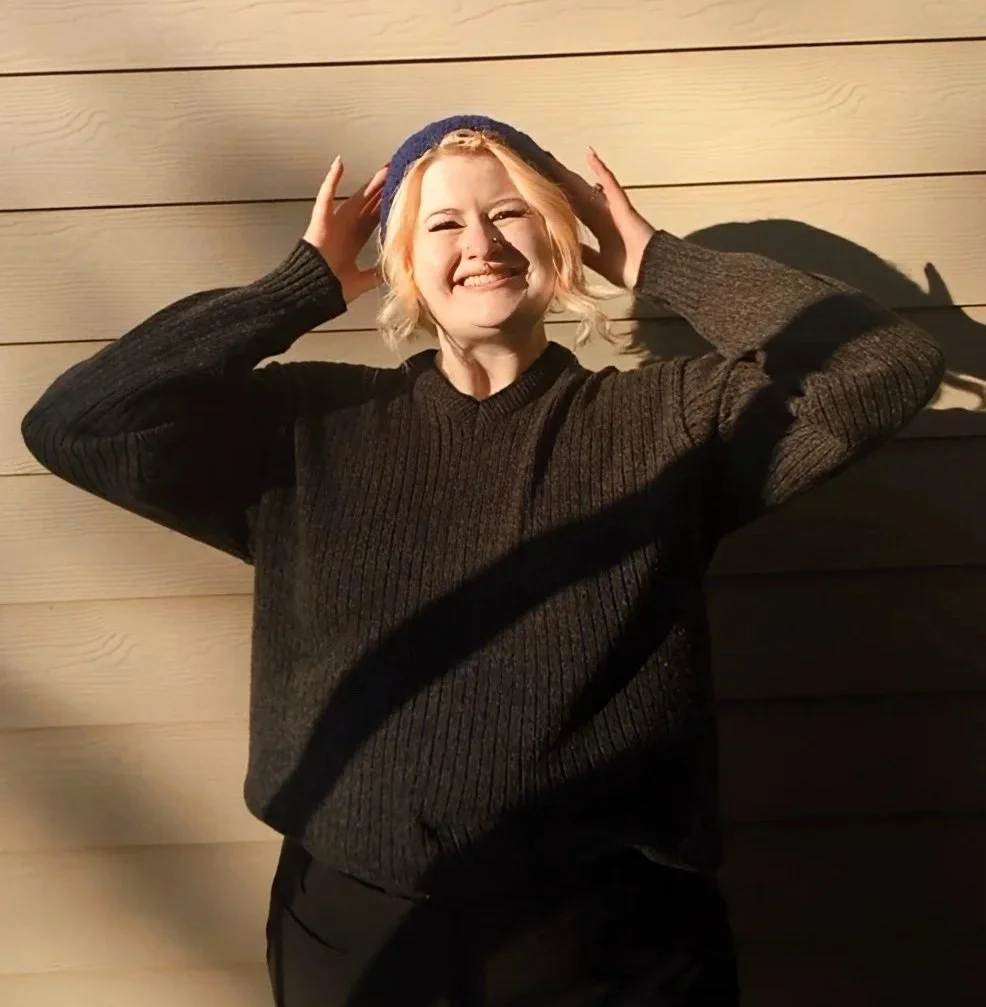 A young woman with blonde hair stands in the sun. They are wearing a black knit sweater, black jeans, and a crochet blue hairband. Their arms are raised, with fingers touching the headband.