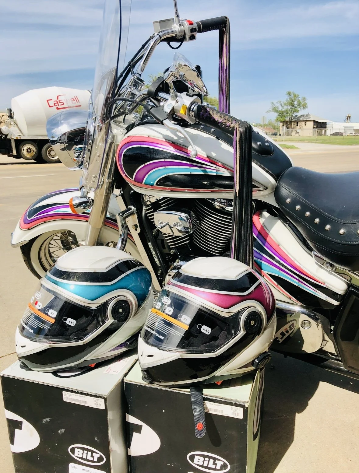 Colorful motorcycle with two motorcycle helmets on boxes in front, helmets are black, blue, and pink with white, on a sunny day with a tanker truck in the background.