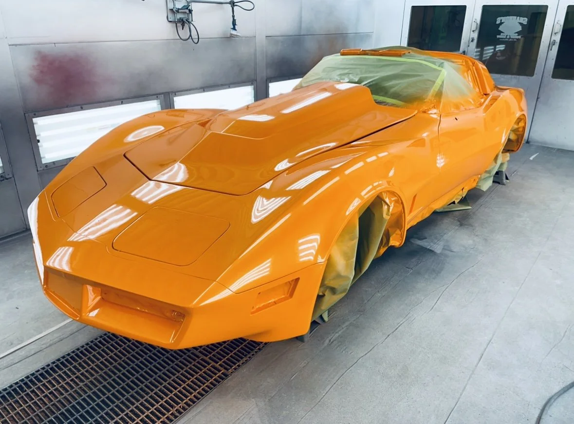 Orange sports car in a paint booth, covered with masking tape and paper for painting.