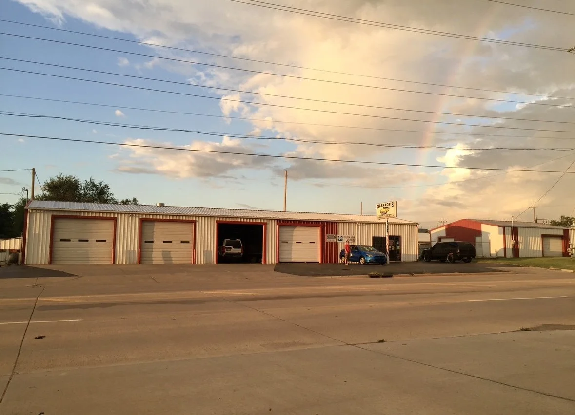 A large metal building with four garage doors, two of which are open with cars inside. There are two cars parked outside, and a person standing near the blue car. The building has a sign for 'Shannon's' and is set against a sky with clouds and a faint rainbow, with power lines overhead and a wide street in the foreground.