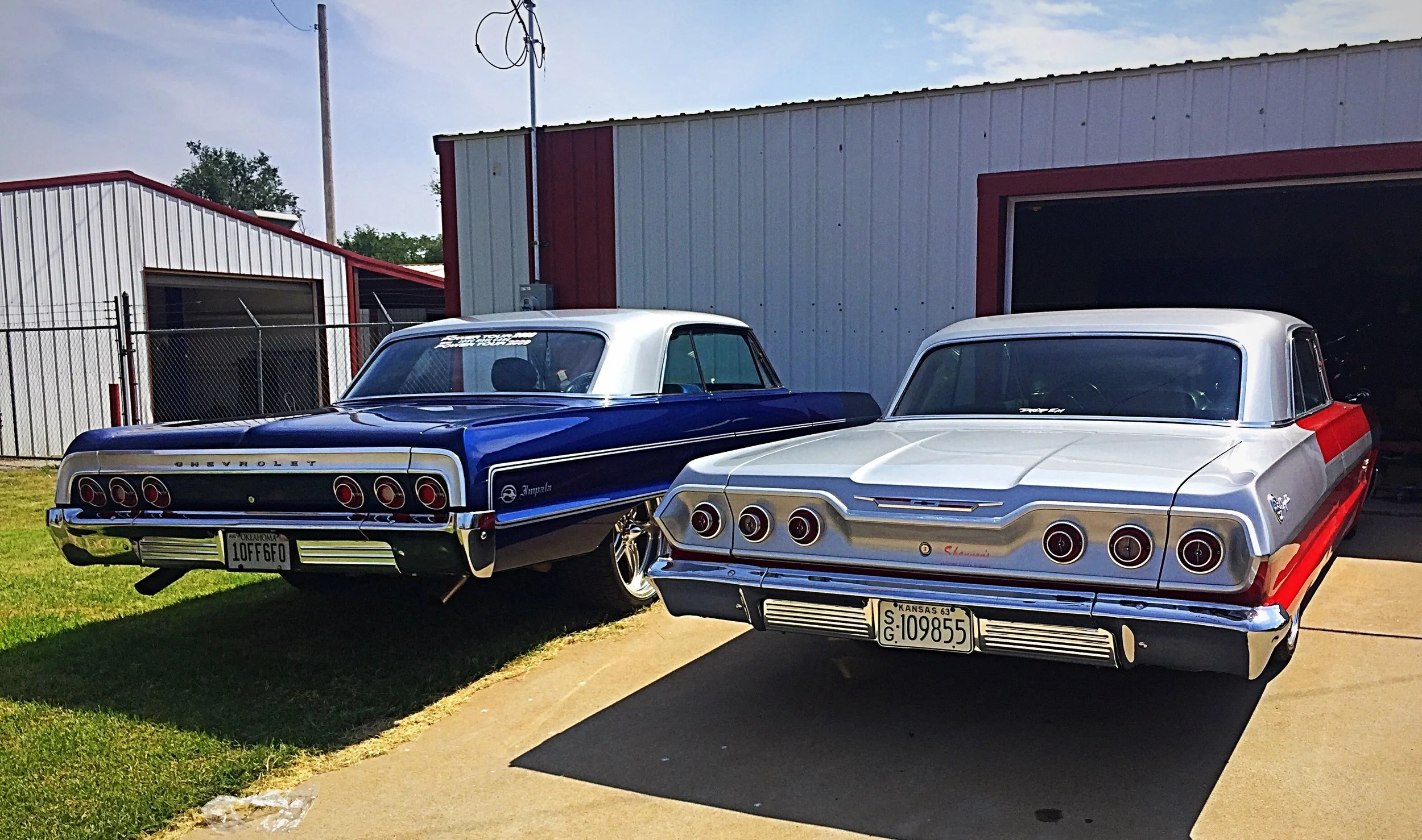 Two classic Chevrolet Impala cars parked outside a building with metal siding, one blue and white, the other light gray with red accents.