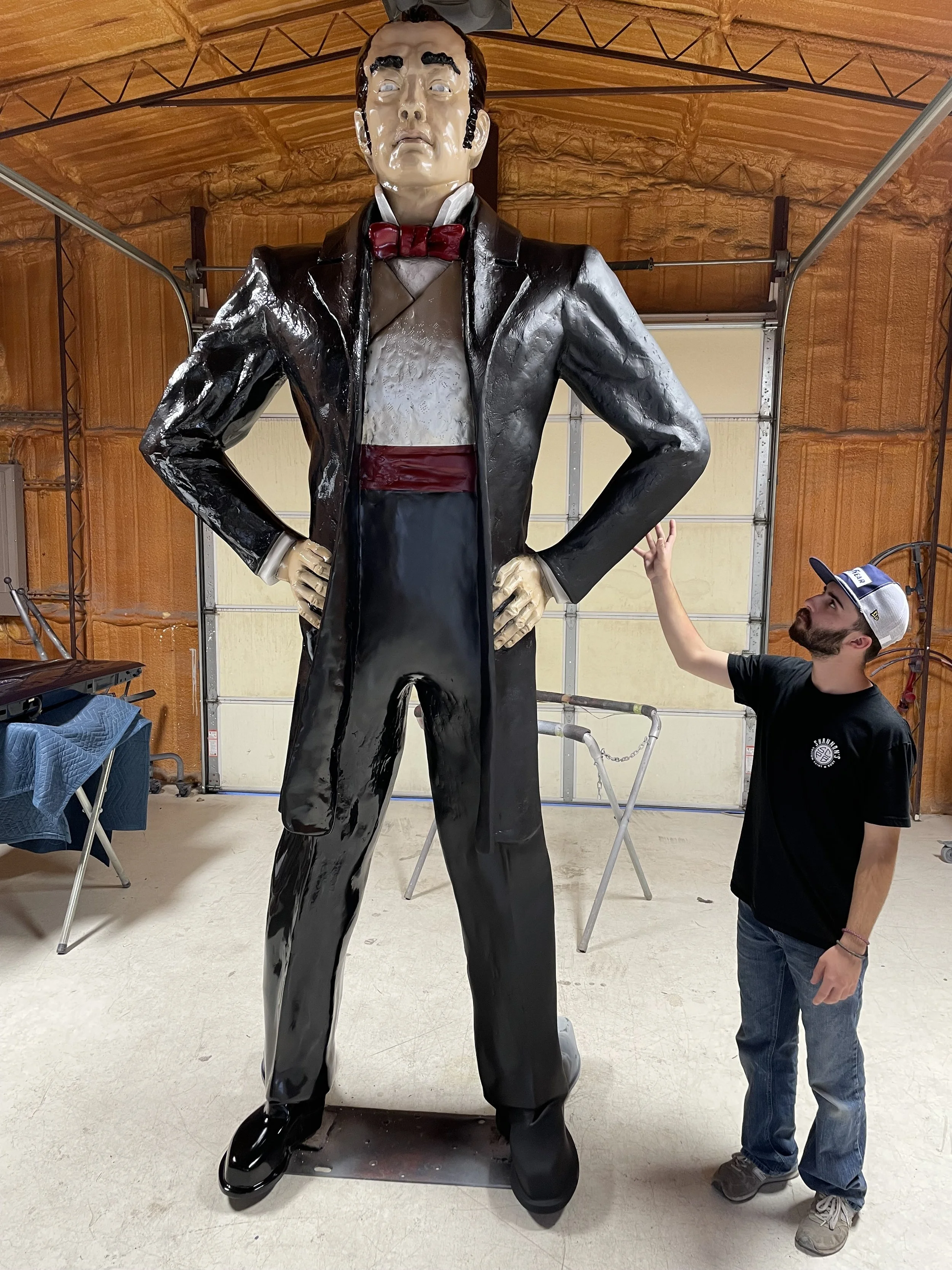 A young man standing next to a 12 ft tall butler statue inside a garage. The man is wearing a black T-shirt, jeans, and a cap, and is reaching up with his right hand to touch the statue's arm.
