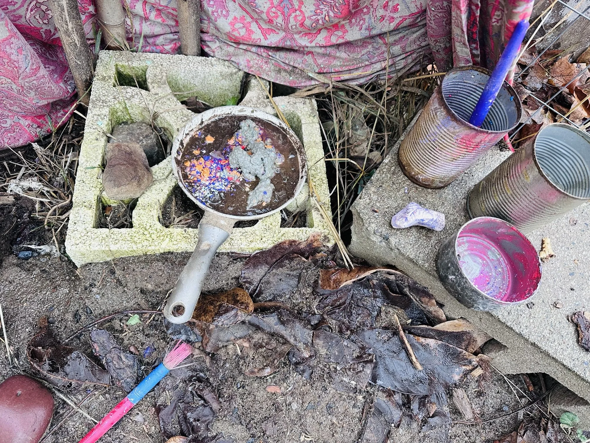 A makeshift outdoor workspace with a paintbrush, three tin cans, a saucepan with water and paint, a concrete block, and fallen leaves, suggesting an art or painting setup in a natural setting.