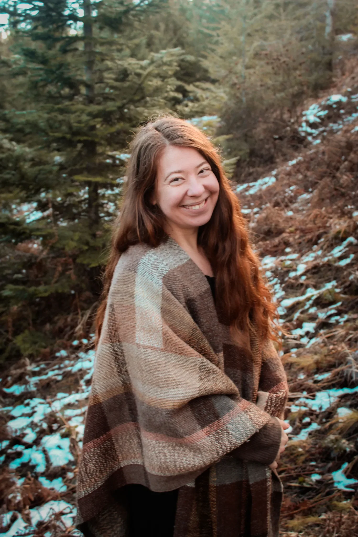 A young woman with long brown hair smiling outdoors in a wooded area. She is wrapped in a brown and beige plaid blanket, with trees and patches of snow on the ground in the background.