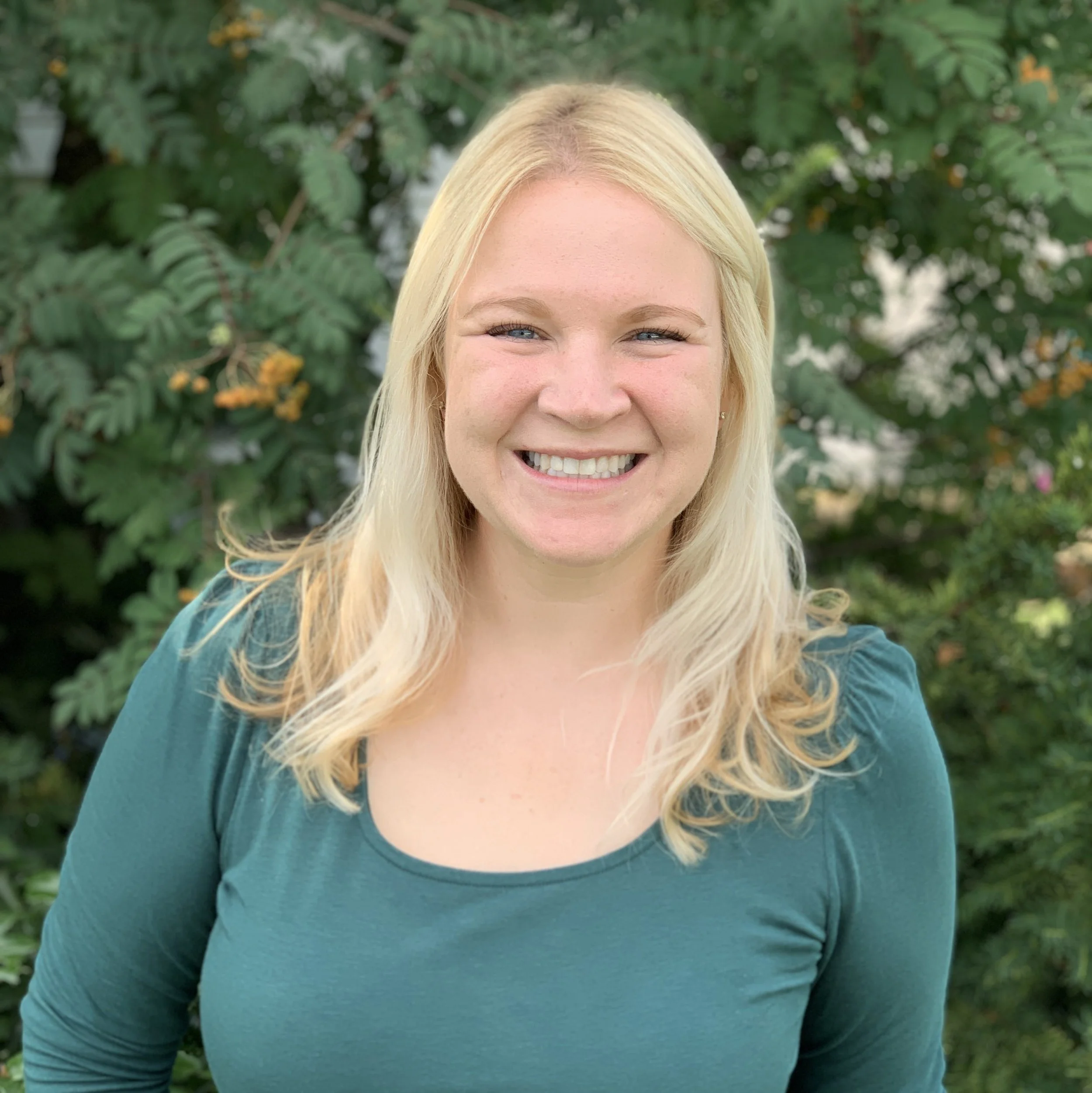A woman with long blonde hair smiling outdoors against green foliage.