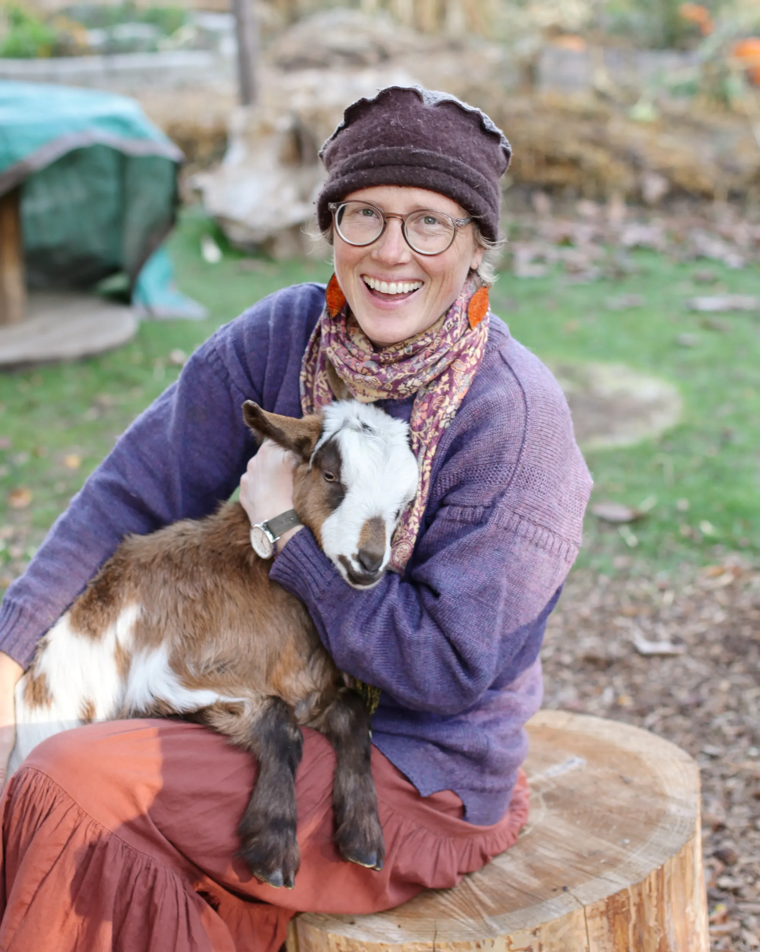A woman sitting on a tree stump outdoors, holding a baby goat, smiling, with trees and a small shed in the background.