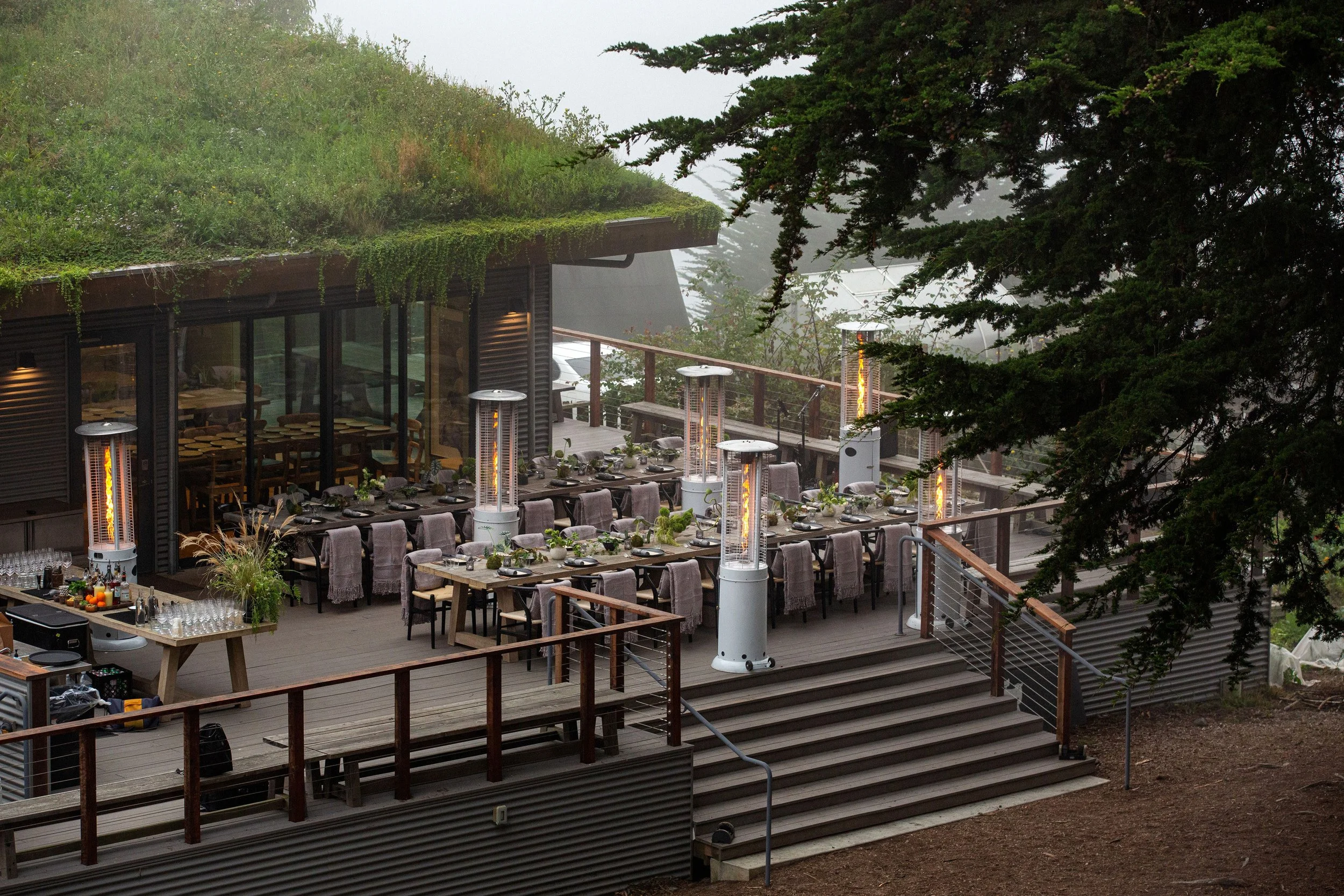 Outdoor restaurant patio with arranged tables set for dining, surrounded by heaters, overlooking a forested area with fog in the background.