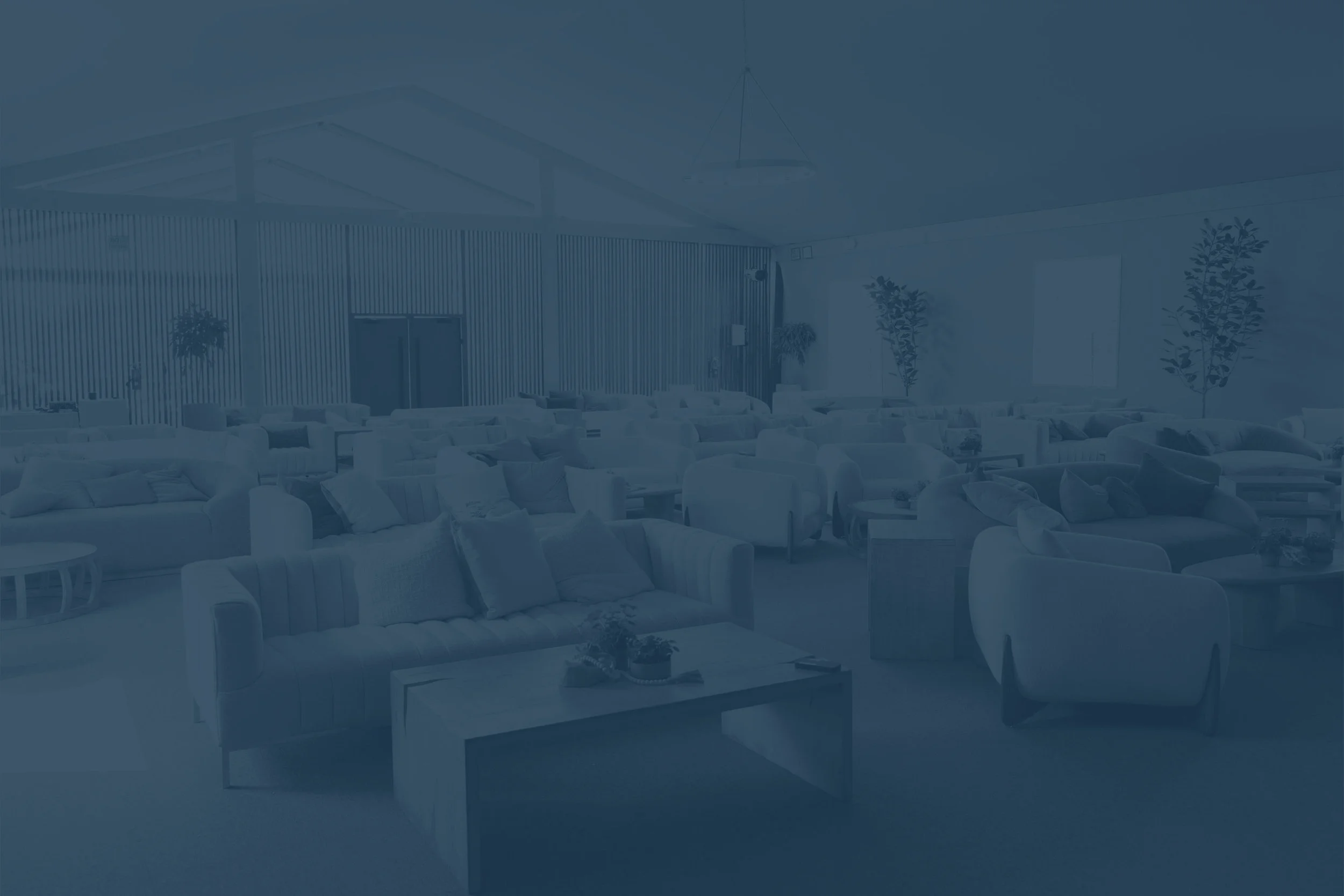 An interior view of a spacious, modern lounge area with many white sofas and armchairs, small tables, potted plants, and decorative elements, with a high ceiling and vertical wooden wall accents.