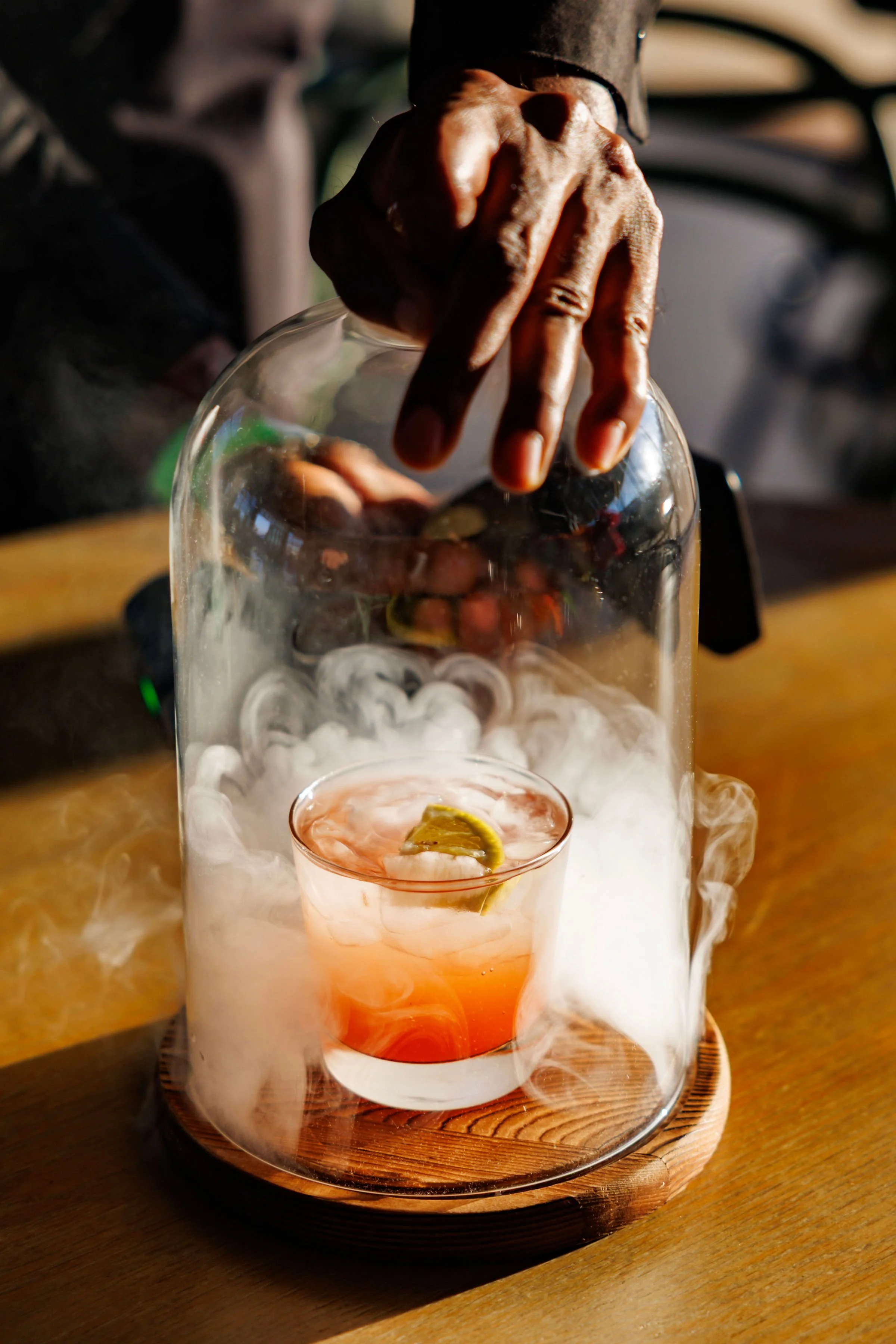 Hand stirring a drink with a slice of lime in a glass, surrounded by aromatic smoke inside a glass container.
