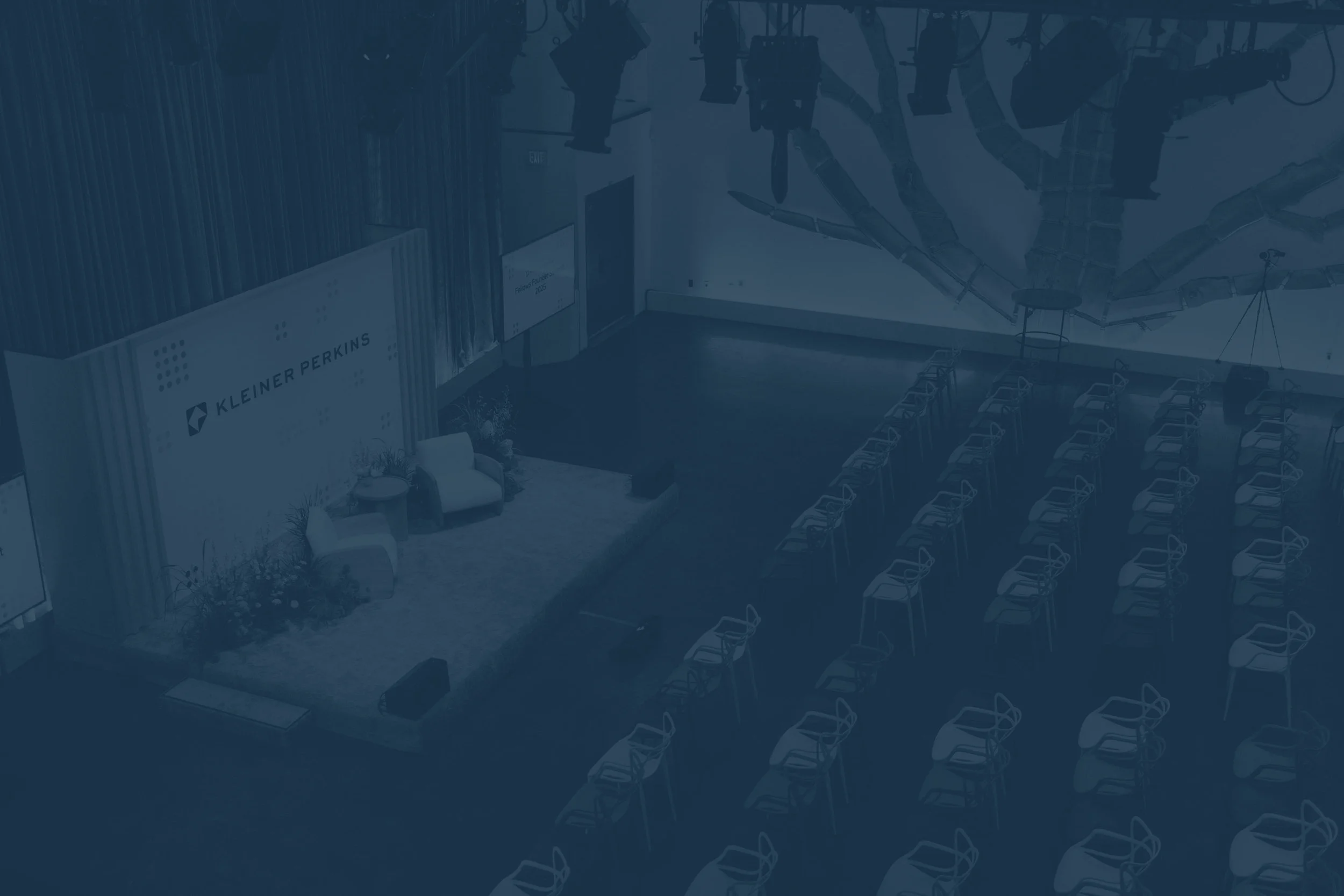 An empty conference room with rows of chairs facing a stage with a screen. The room has a large wall mural, and there is a seating area with chairs and a small table near the stage. The environment appears to be set up for a presentation or event.