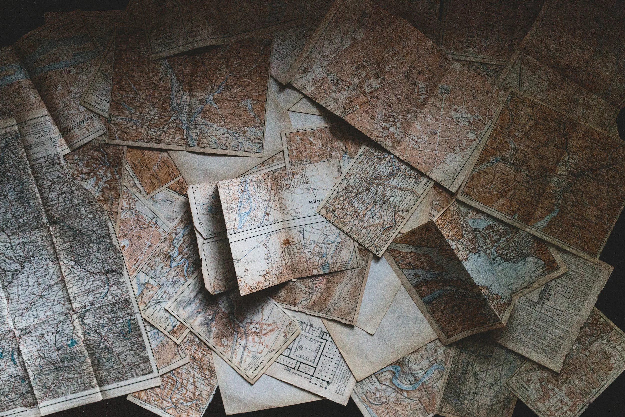 A collection of vintage paper maps of various regions, spread out and overlapping on a surface. The maps show topographical details, roads, rivers, and place names.