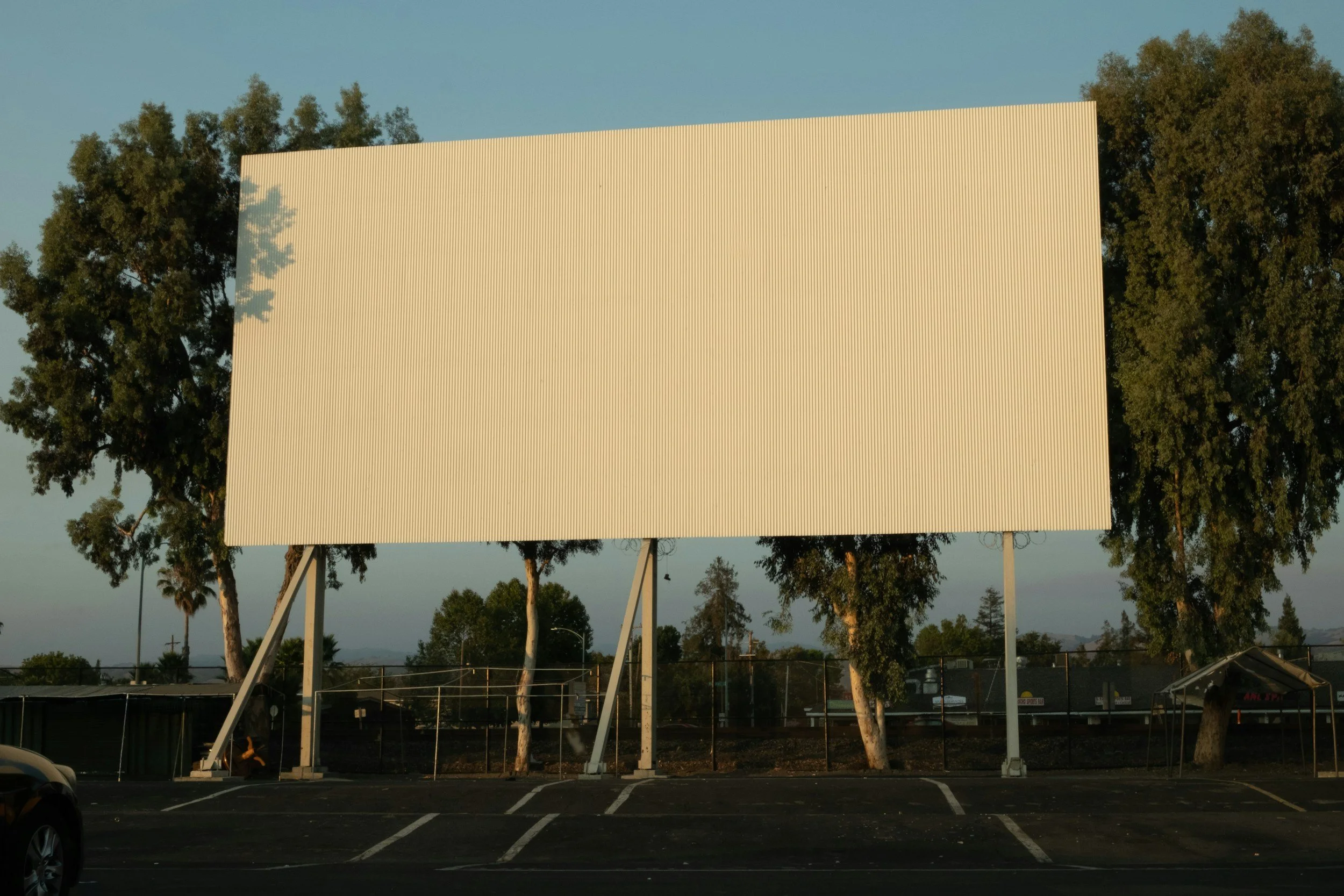 Empty large outdoor bill board on a metal stand with trees and parking lot in the background.