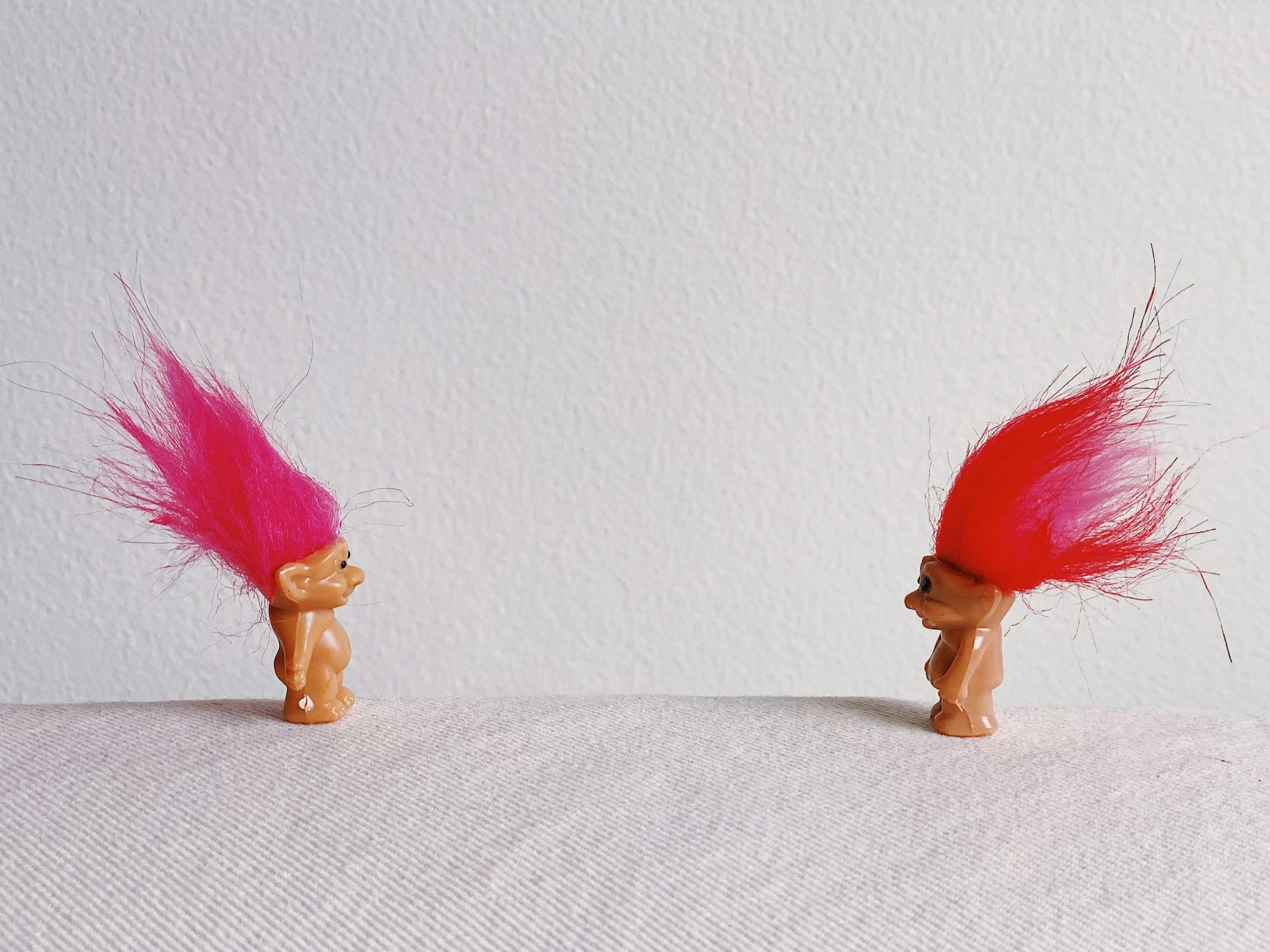 Two small toy figures with bright pink and red hair facing each other on a flat surface against a white wall.