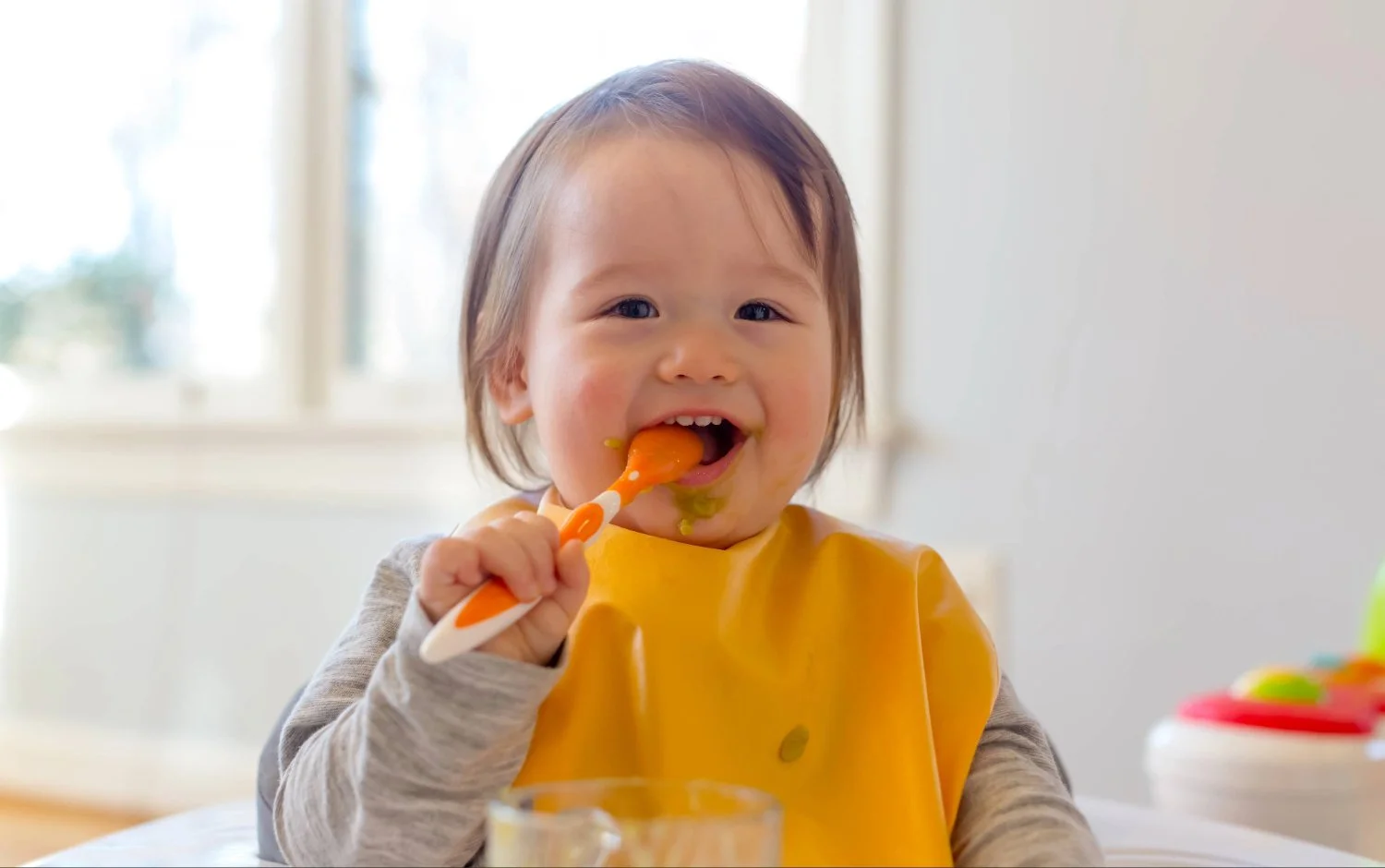 A smiling toddler with brown hair eating food with an orange and white spoon, wearing a yellow bib, working on transitioning to solids during feeding therapy in New York City.