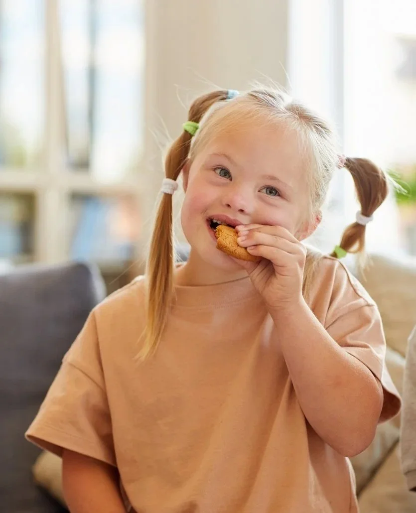 A young girl with blonde hair in pigtails eating a cookie indoors, smiling at the camera, working on chewing and swallowing during feeding therapy for kids in New York City.