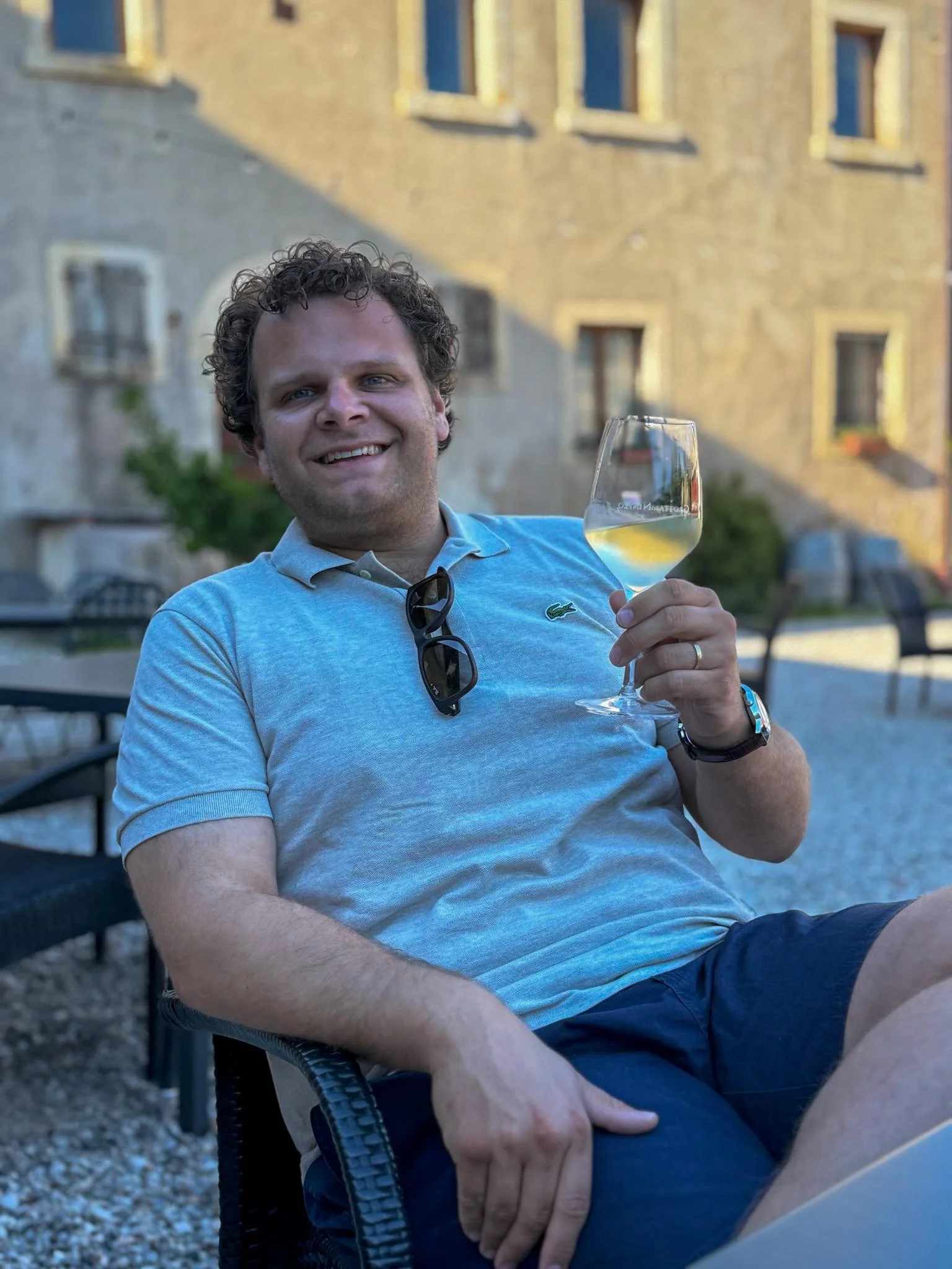 A man smiling and sitting outdoors, holding a glass of white wine, wearing a light gray polo shirt with sunglasses hanging on his shirt collar, and a wristwatch, with a building and chairs in the background.
