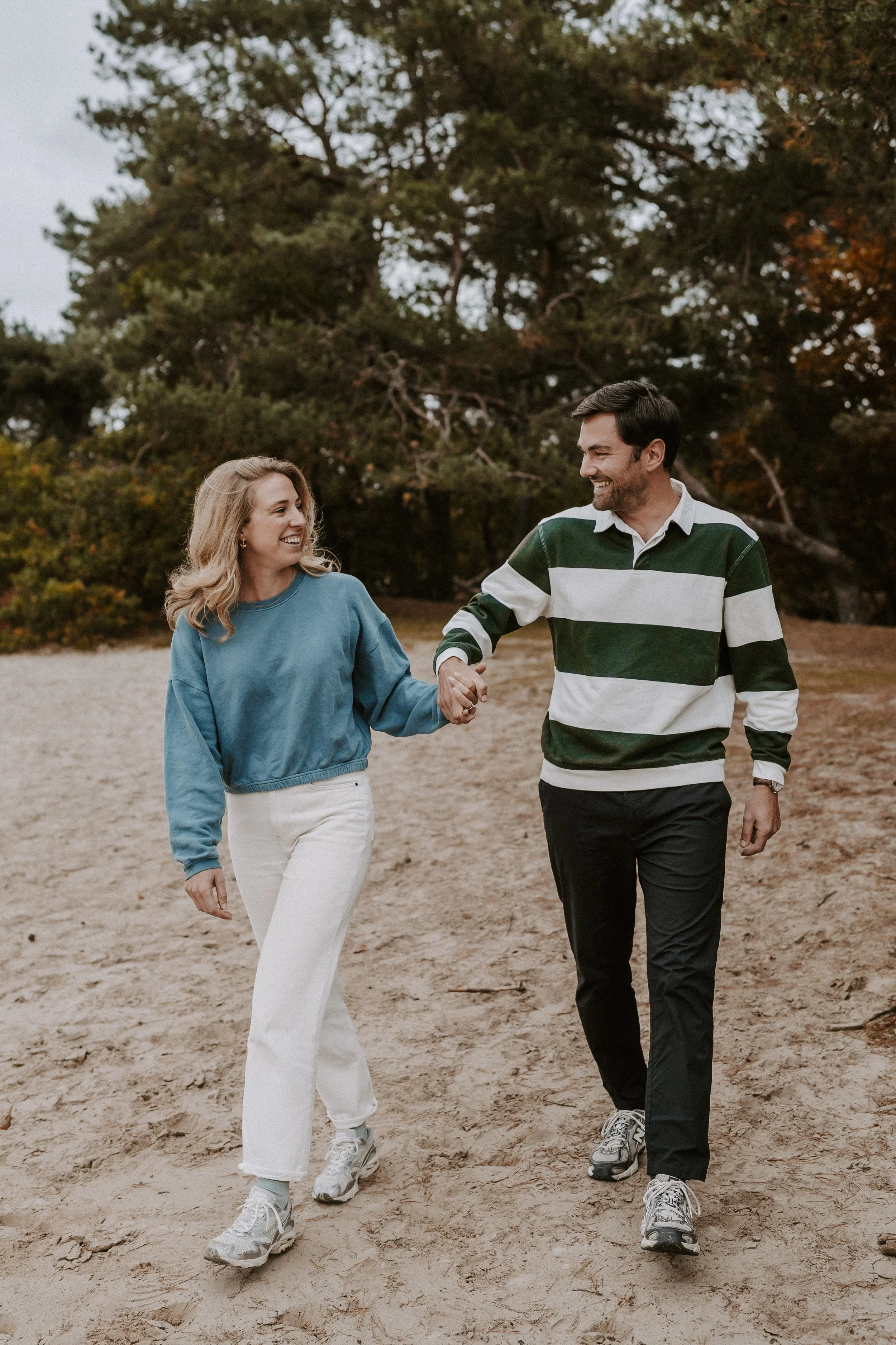A smiling woman and man walking hand in hand outdoors on a sandy path with trees in the background.