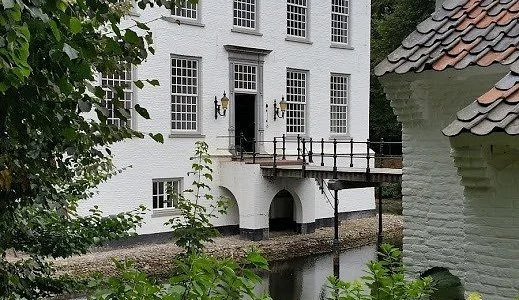 White historic building with multiple windows, a small bridge, and a waterway, surrounded by greenery and a small white structure with a tiled roof.