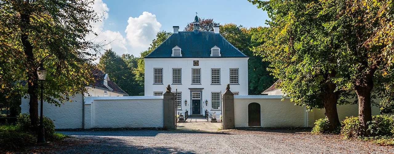 A large white historic building with multiple windows and a dark roof, surrounded by trees, with a gravel driveway leading up to the entrance.