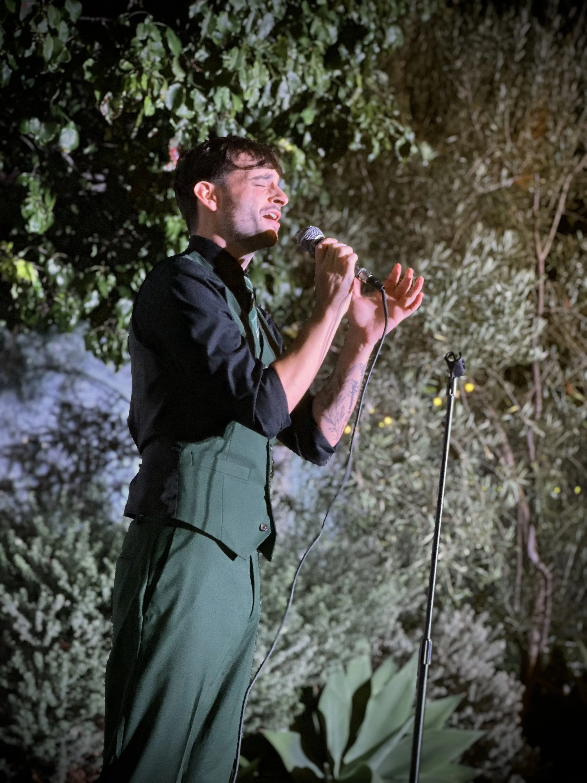 A singer with dark hair, wearing a black shirt and green apron, performs with a microphone on a stage with green plants and bushes background.