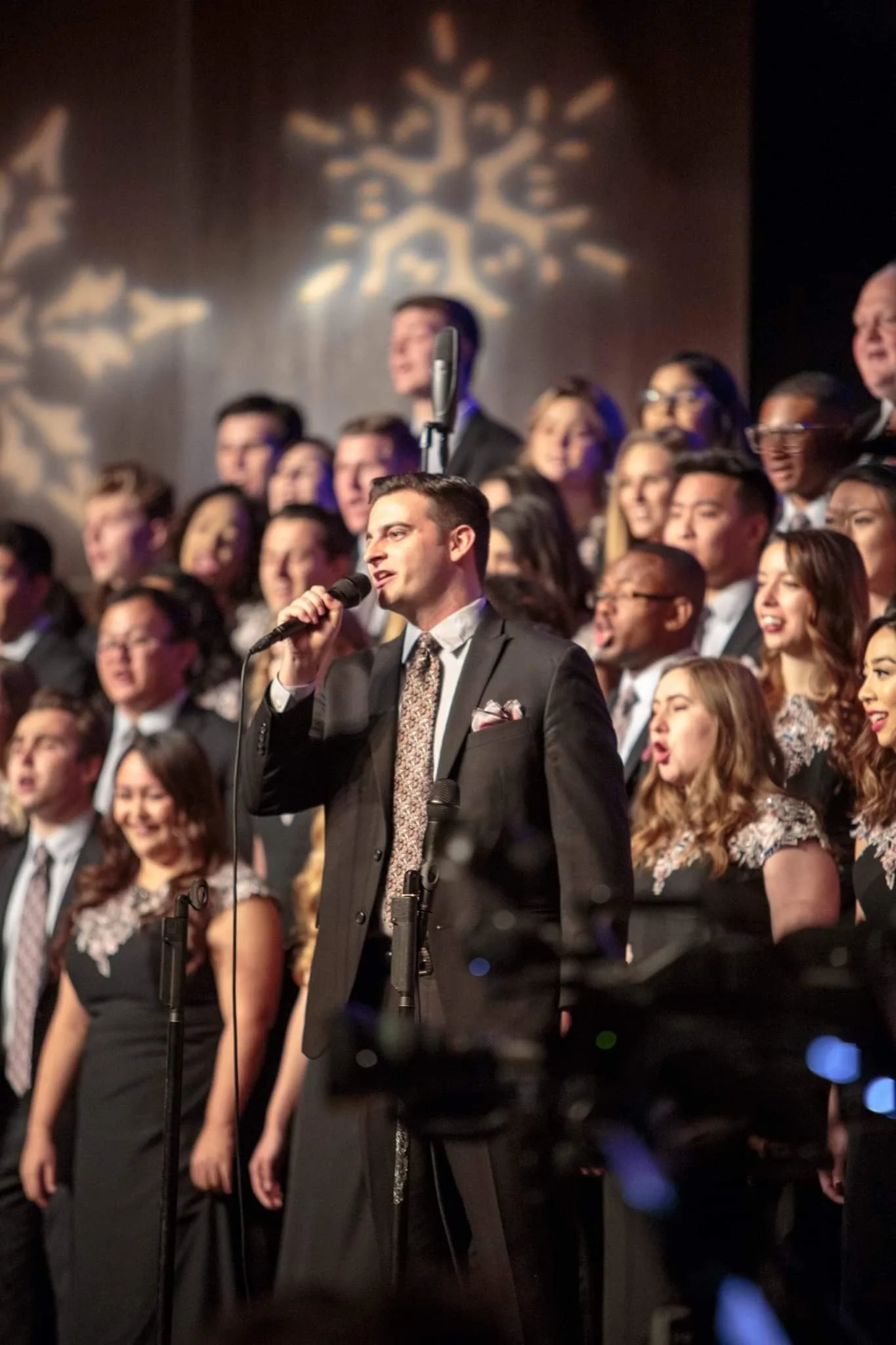 A male choir conductor or soloist in a dark suit holding a microphone while surrounded by a choir of men and women in formal black and floral dresses, performing in a concert hall with snowflake-shaped light projections on the wall.