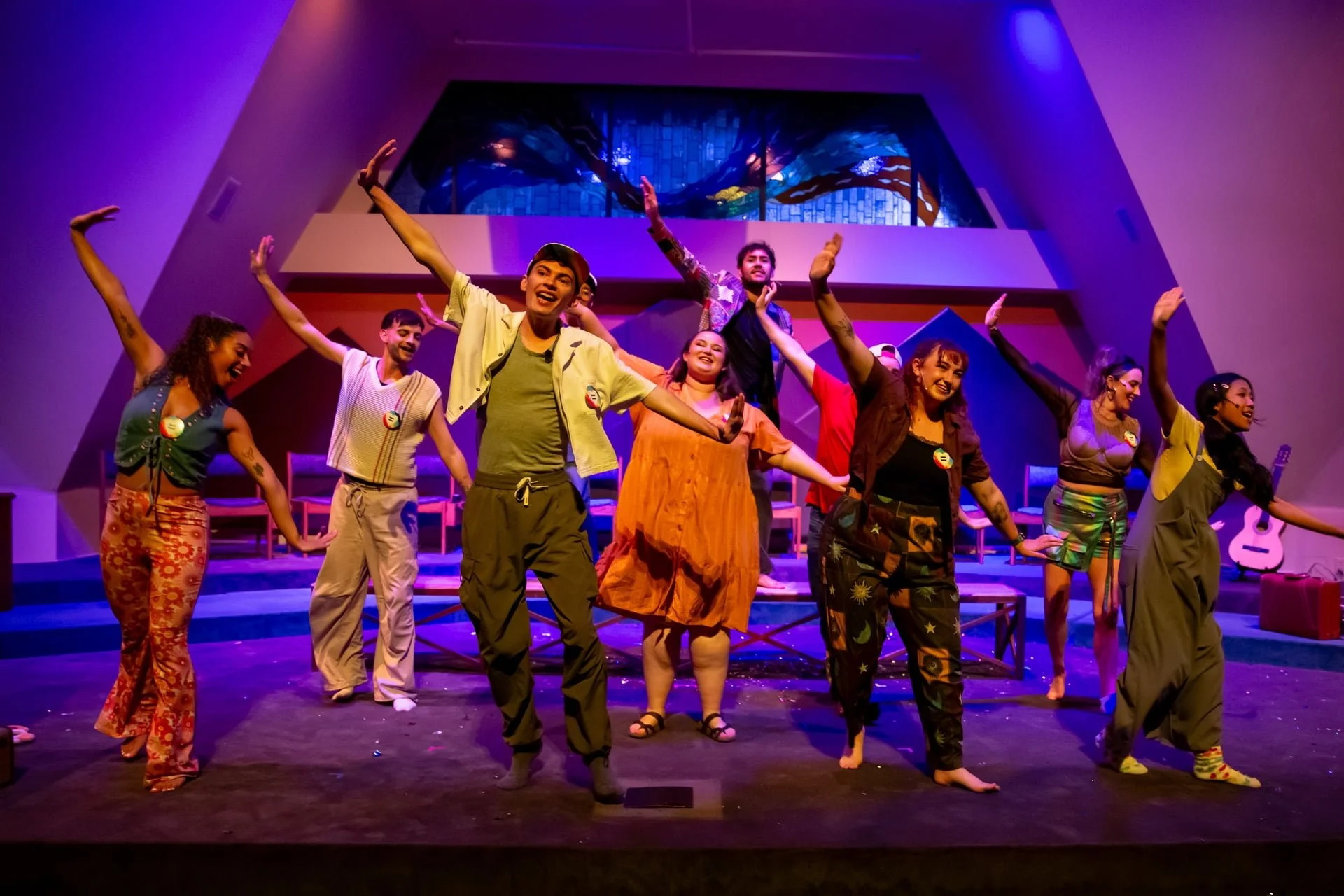 A group of nine people performing on a brightly lit stage with colorful lighting, dressed in casual and colorful clothing, smiling and holding hands, with a musical instrument visible in the background.
