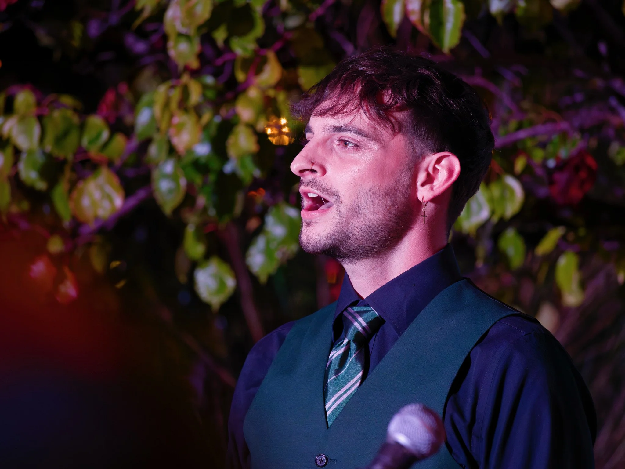 A young man singing or speaking into a microphone outdoors at night, wearing a dark shirt, vest, and striped tie, with a green earring and a nose ring, background of leafy shrubbery with colorful lighting.