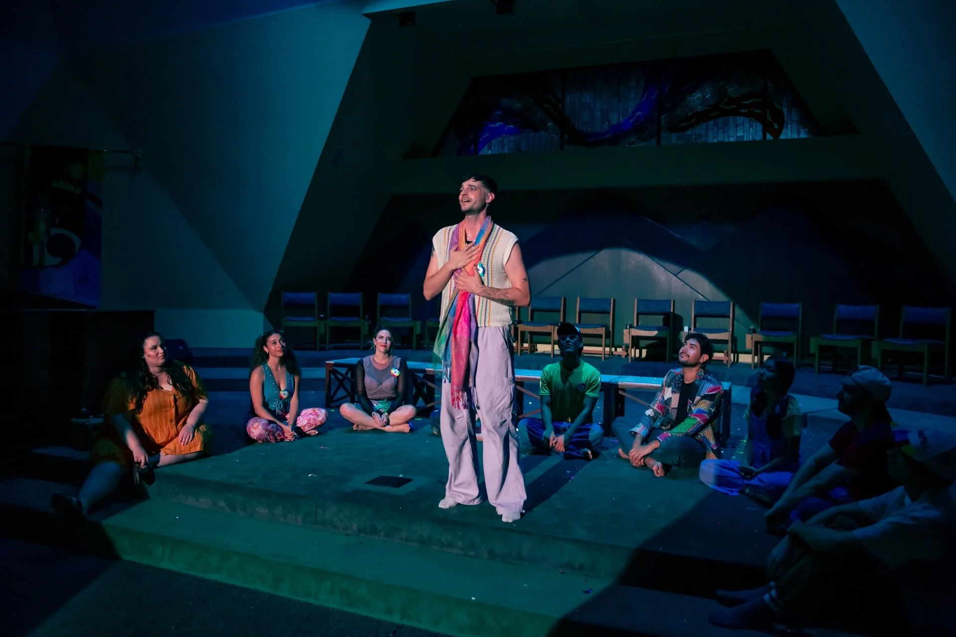 A group of people sitting on the stage in a semi-circle while a man stands in the center, speaking or singing with his hand on his chest. The scene appears to be a theatrical or community performance inside a concert hall or auditorium.