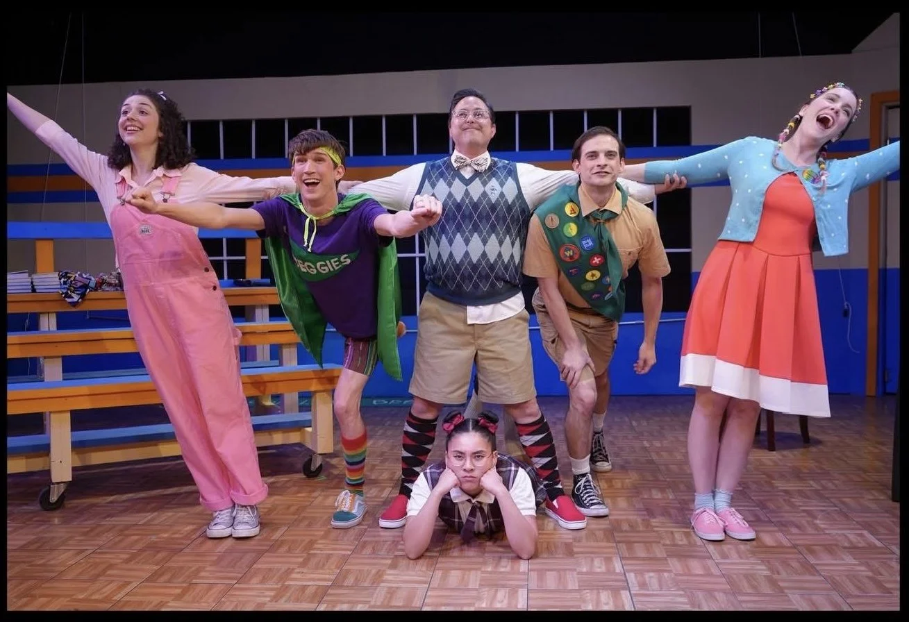 Group of six young actors on stage performing a play, with four standing and two sitting or lying on the floor, in colorful costumes and expressive poses.