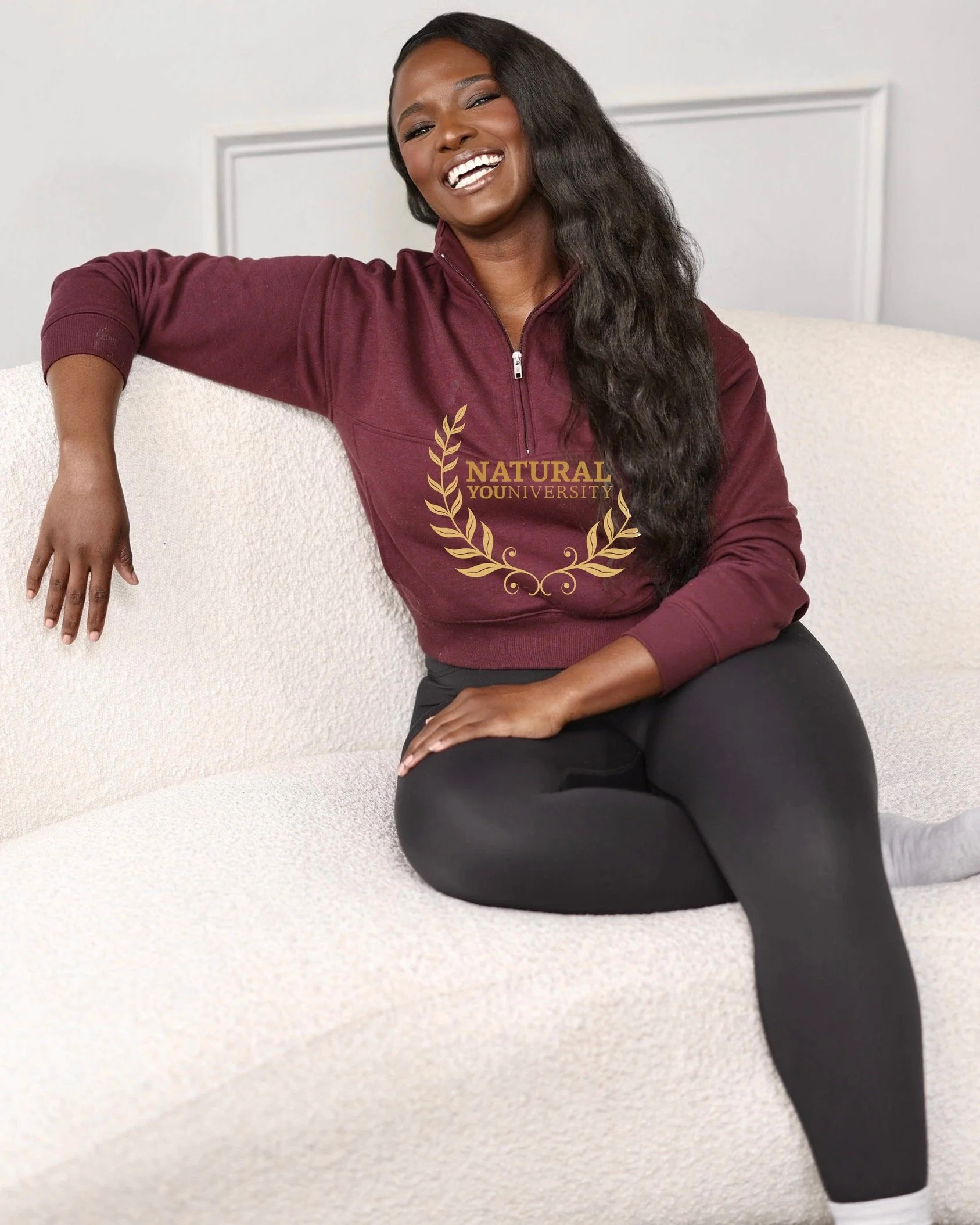 A smiling woman sitting on a cream-colored couch, wearing a maroon zip-up sweatshirt with the logo 'Natural Youniversity' and black leggings.