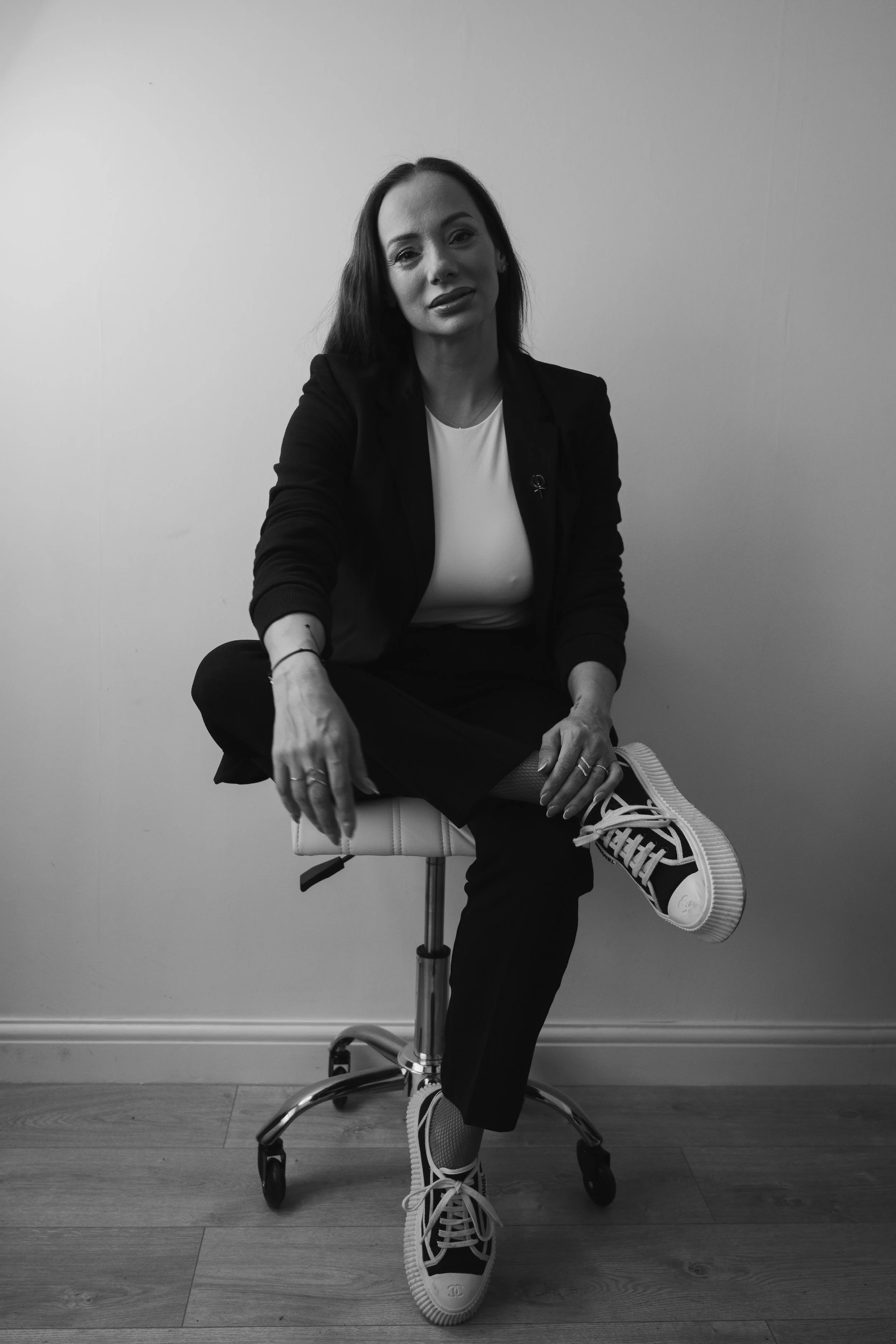 Black and white photo of a woman sitting on a swivel chair with one leg crossed over the other, wearing sneakers, a white top, and a dark blazer, in a minimal indoor setting.