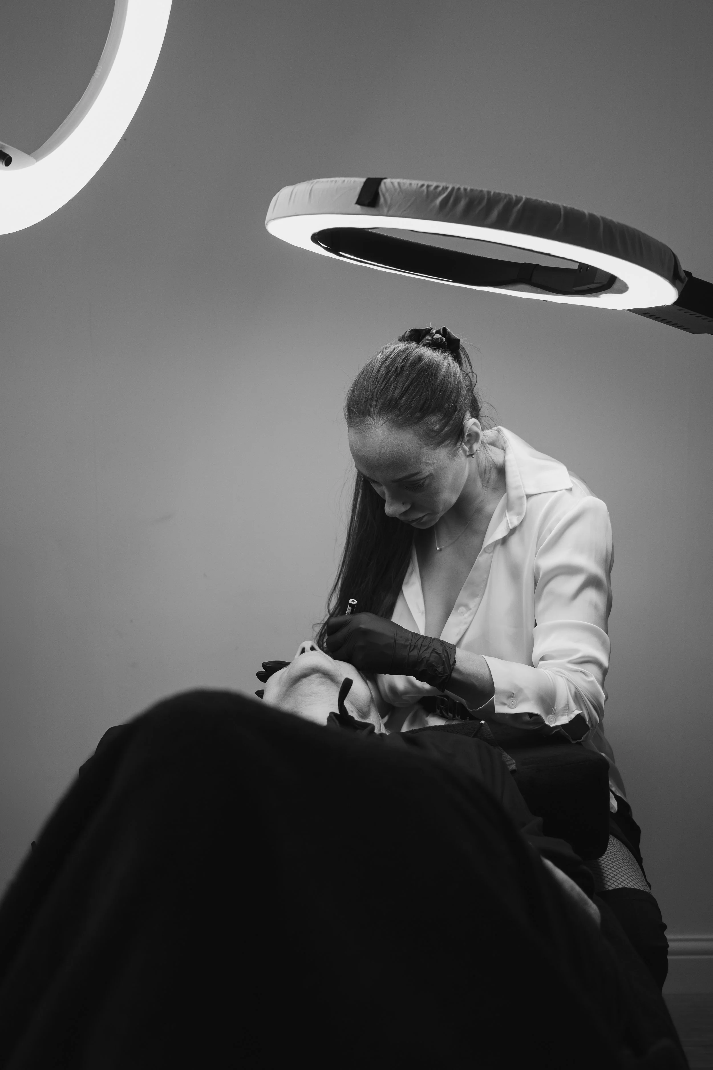 A woman performing a cosmetic procedure on a client in a clinical setting, with overhead lighting.