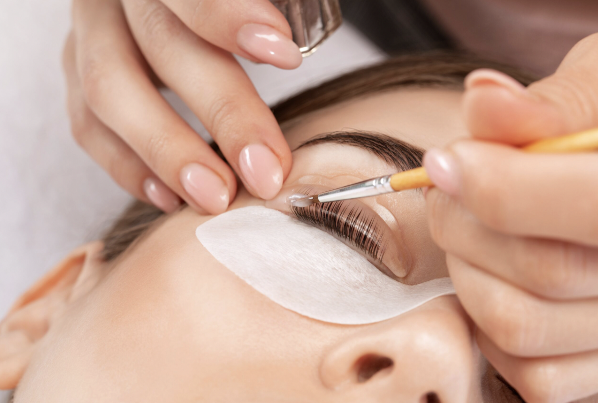 A person receiving an eyelash extension treatment, with a technician applying adhesive to individual eyelashes while the client's eye is covered with a protective pad.