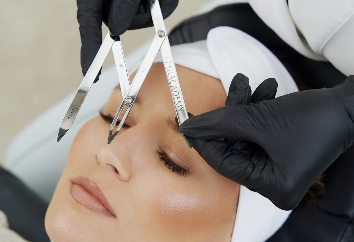 A person receiving an eyebrow tattooing procedure, with a technician using a tattooing tool and calipers labeled 'Phibrows' on the person's eyebrow.