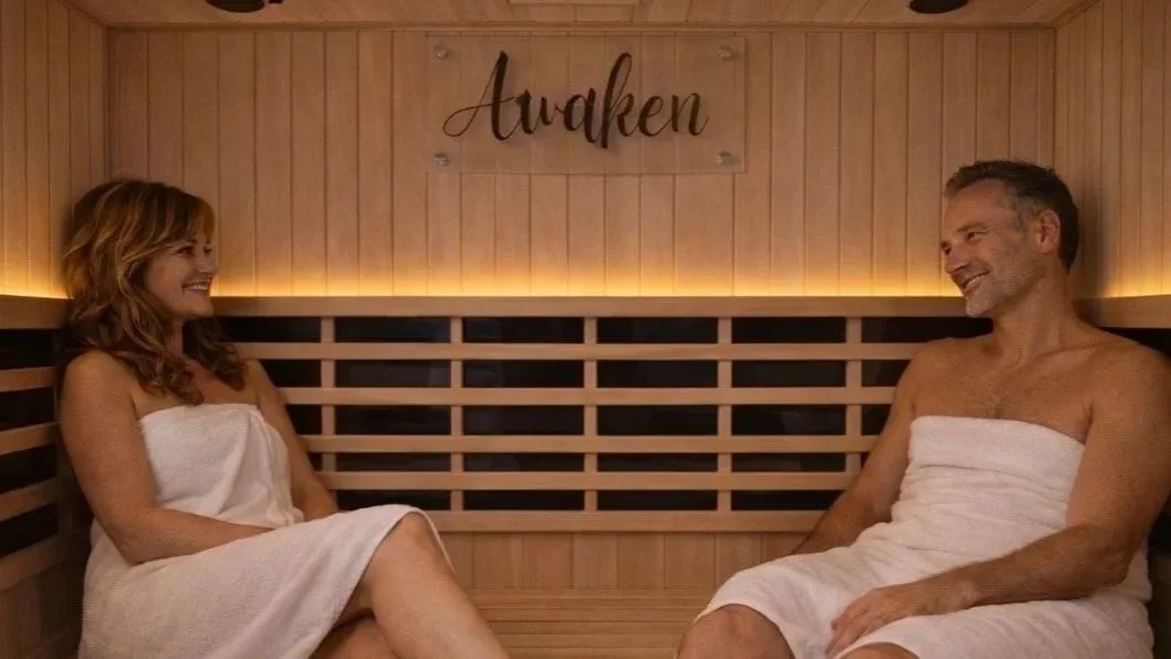 A man and woman sitting in a sauna, wrapped in white towels, smiling at each other, with the word 'Awaken' on the wall behind them.