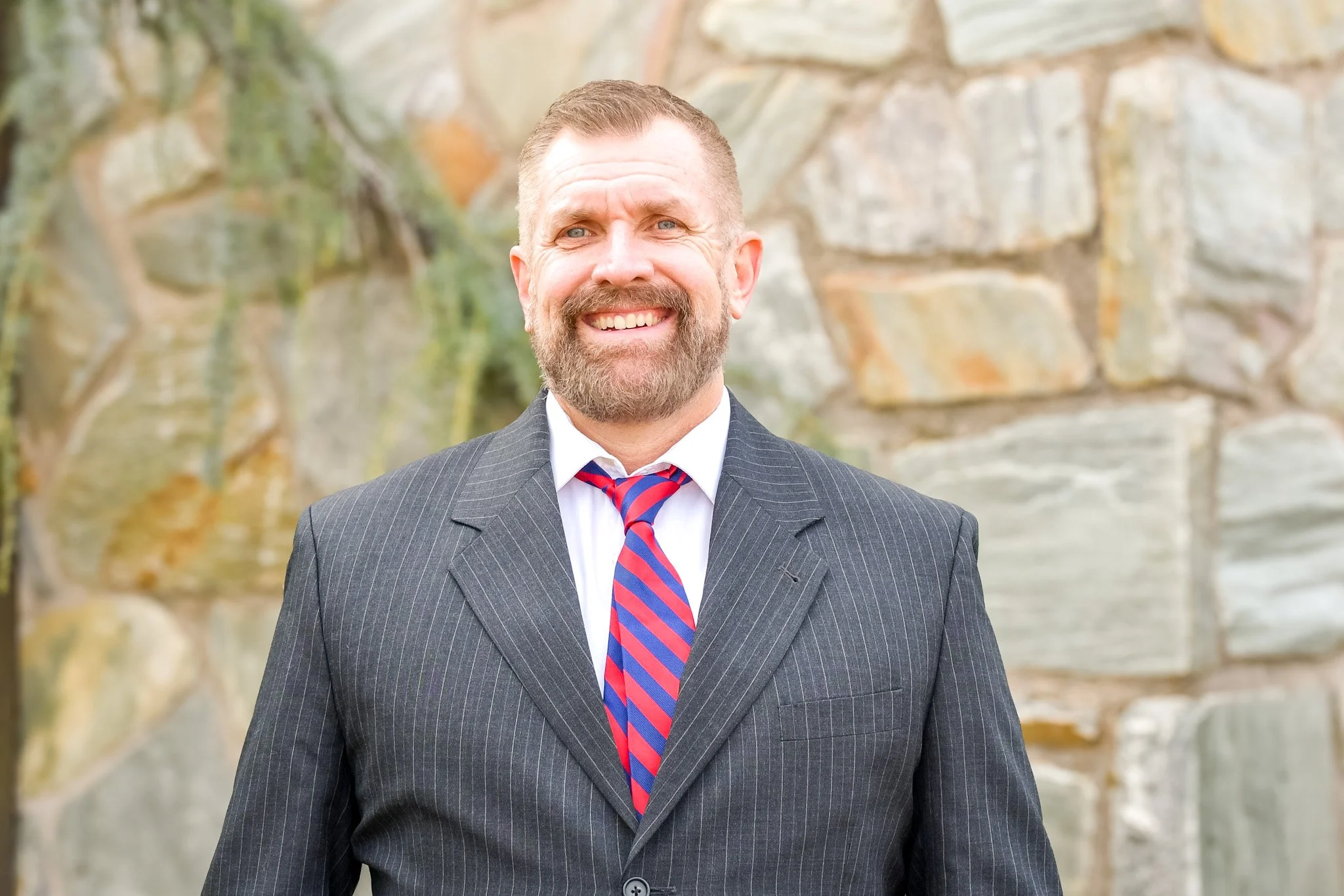 Bill Griffin with a beard and short hair, wearing a dark pinstripe suit, white shirt, and a red and blue striped tie, standing outdoors in front of a stone wall.