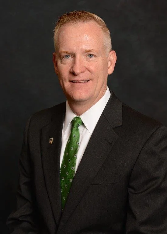 A man with light skin, short, blond hair, wearing a black suit, white shirt, and green tie, posing against a dark background.