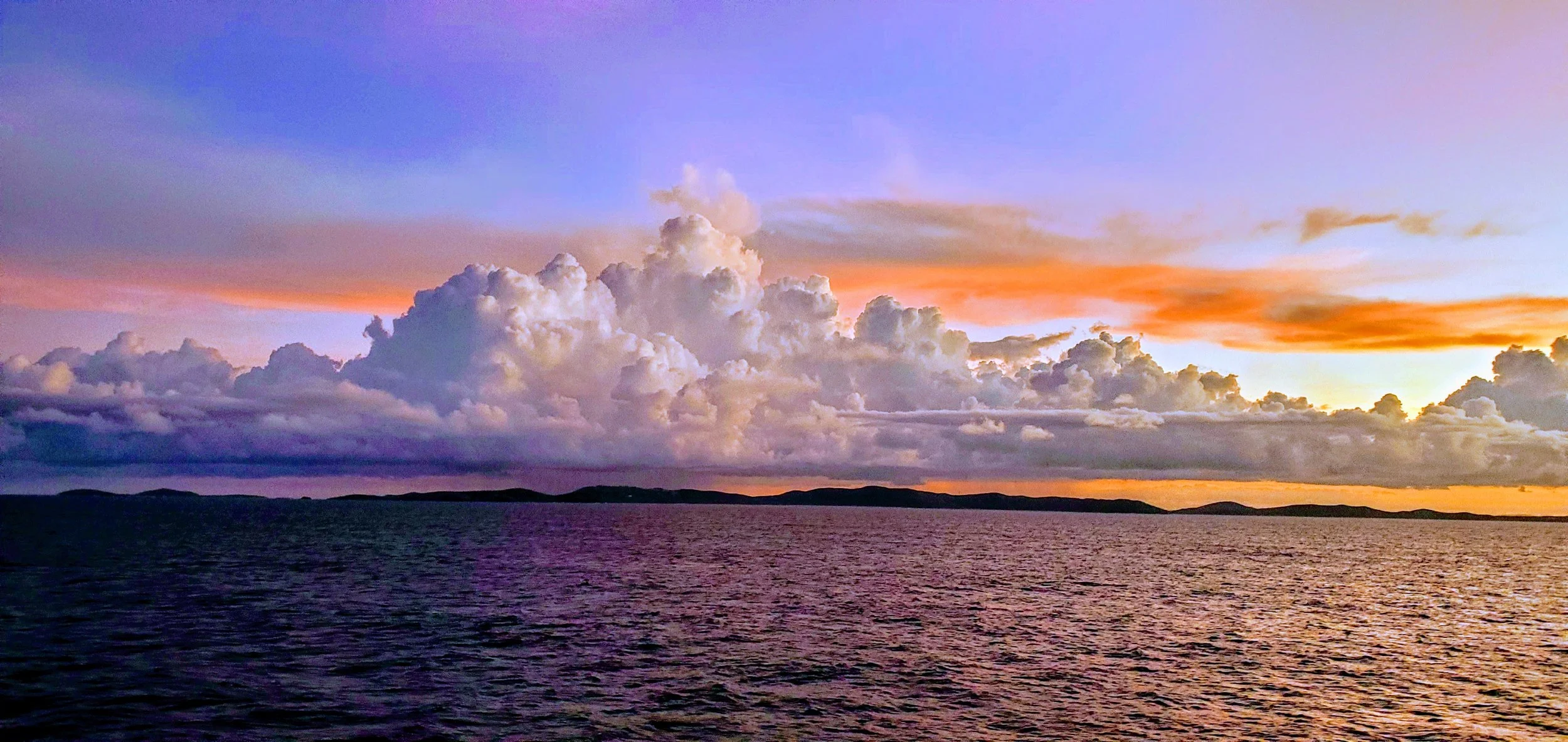 Sunset over calm ocean with distant hills and a colorful sky with clouds.