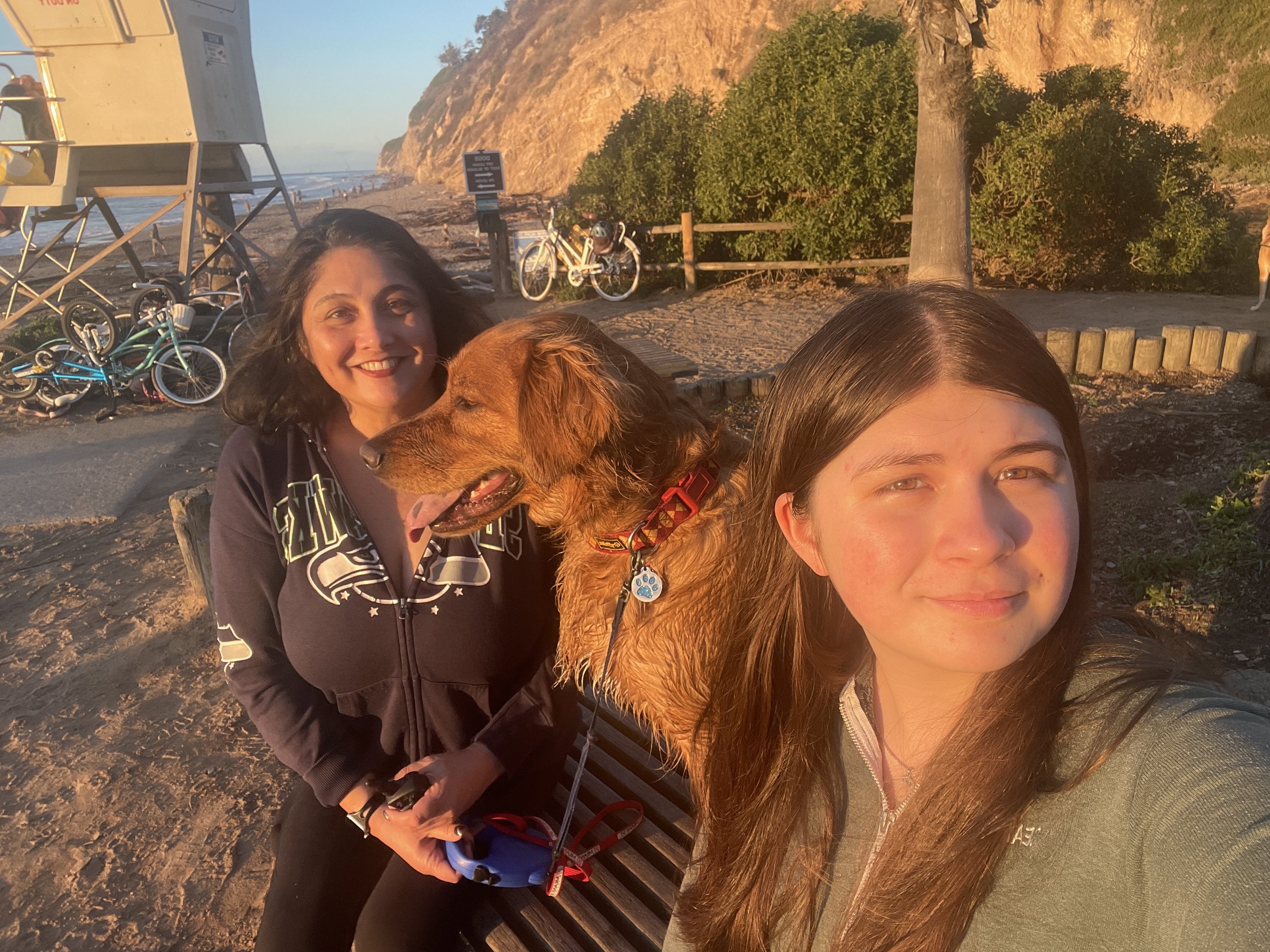 Two women and a dog taking a selfie on a bench at the beach during sunset. There are bicycles and a lifeguard tower in the background, along with trees and cliffs.