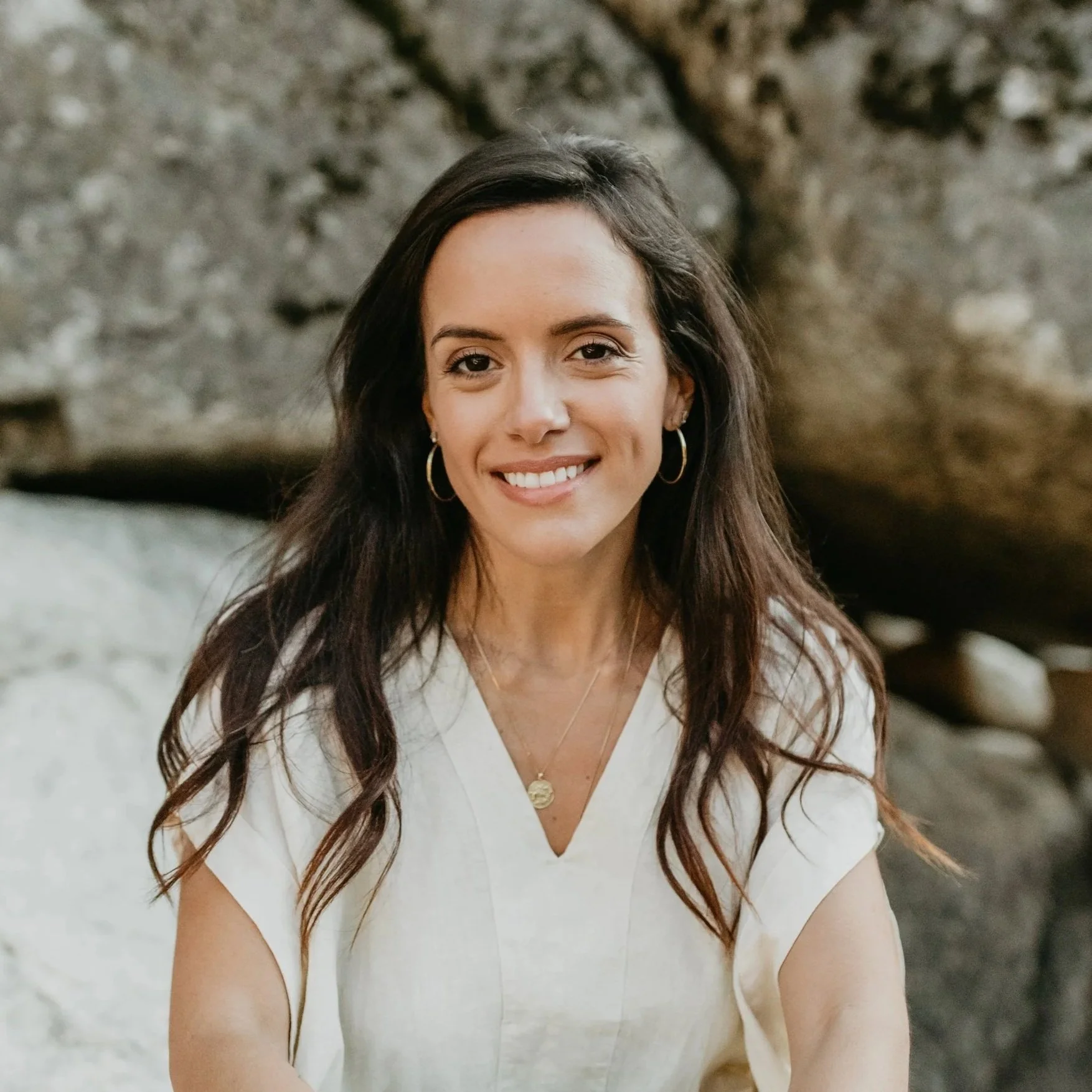 A woman with long dark hair wearing a white top, gold hoop earrings, and layered necklaces, smiling outdoors near rocks and a tree.